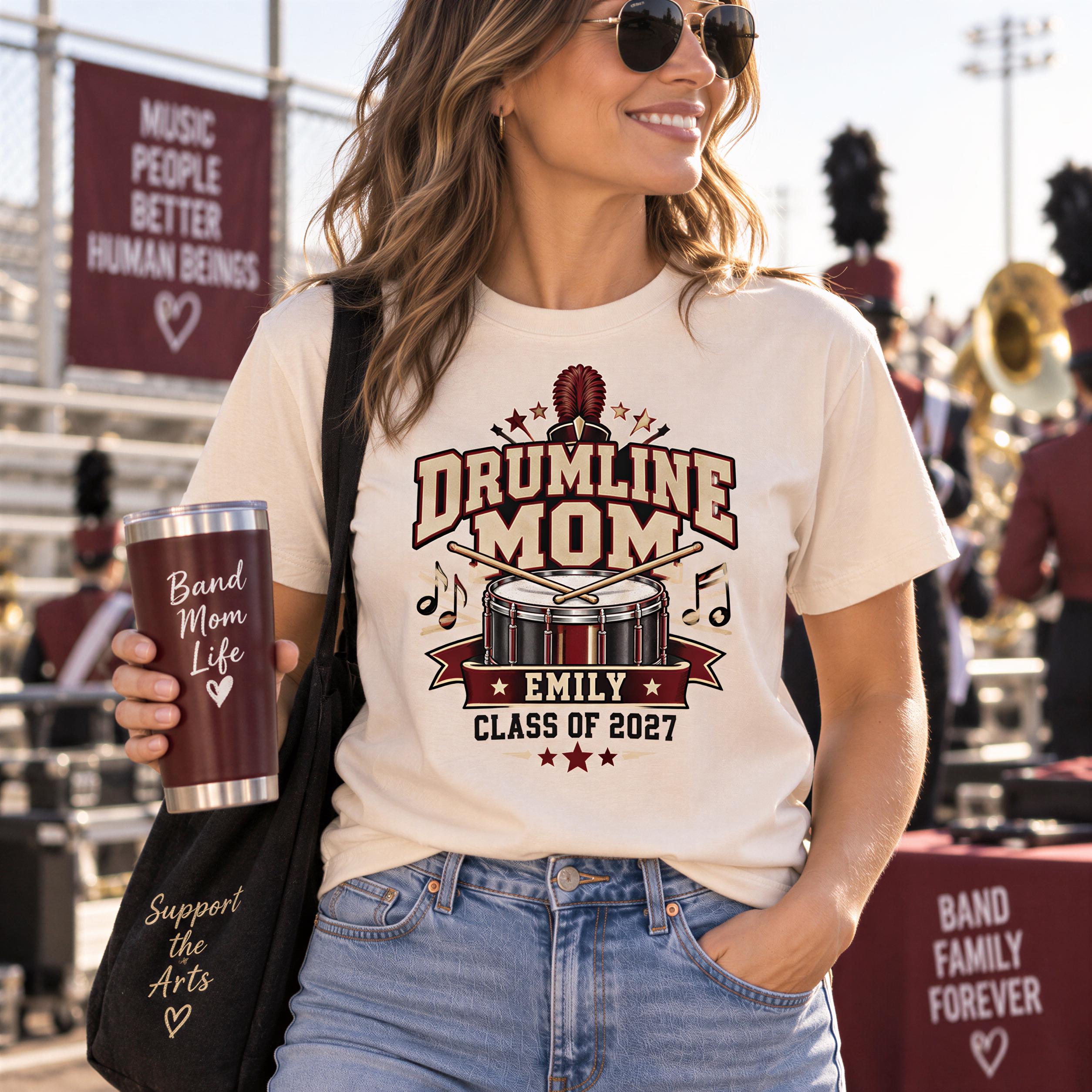 A woman wearing a white t-shirt with a drumline logo and holding a red cup with the text "Band Mom Life" is smiling and standing in front of a marching band.