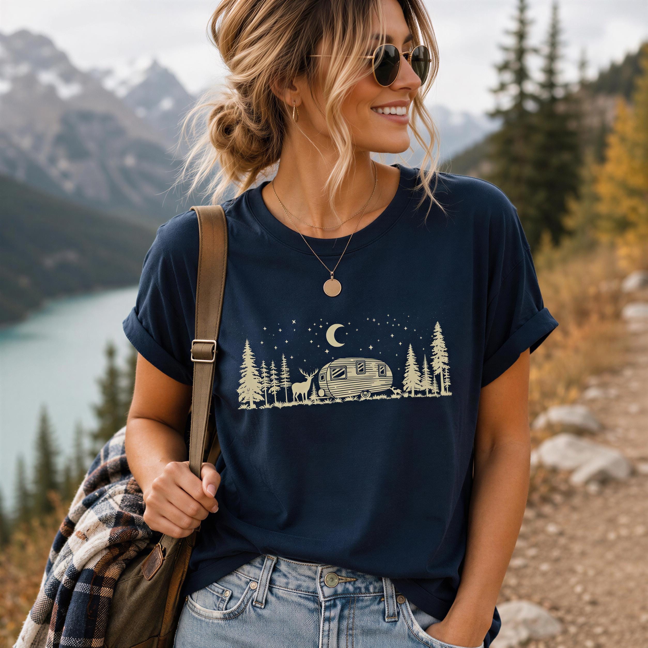 A woman wearing a blue t-shirt with a graphic design of a camper van and a night sky with stars, standing on a dirt path with mountains in the background.
