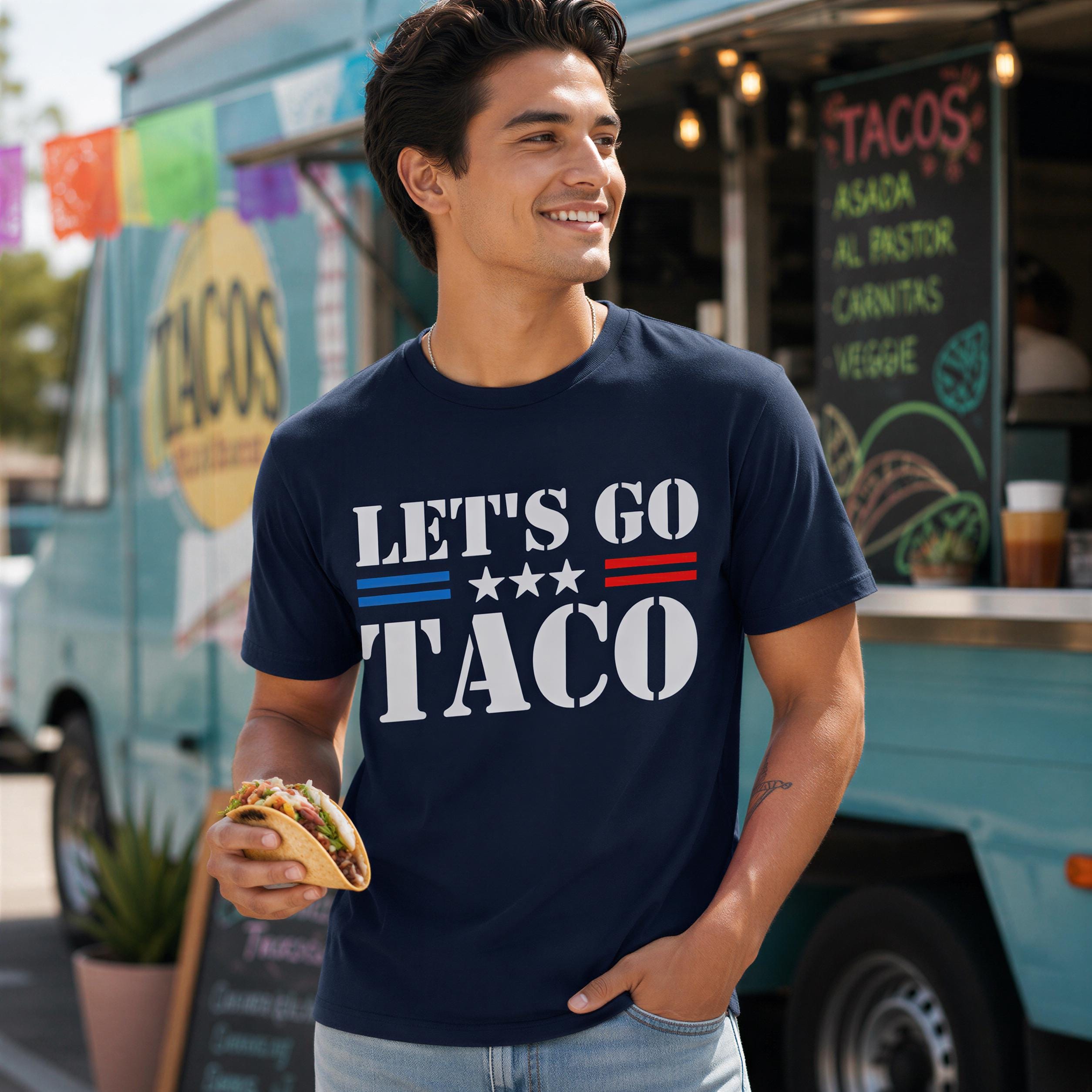 A man wearing a navy blue t-shirt with the text "LET'S GO TACO" is standing in front of a food truck, holding a taco.