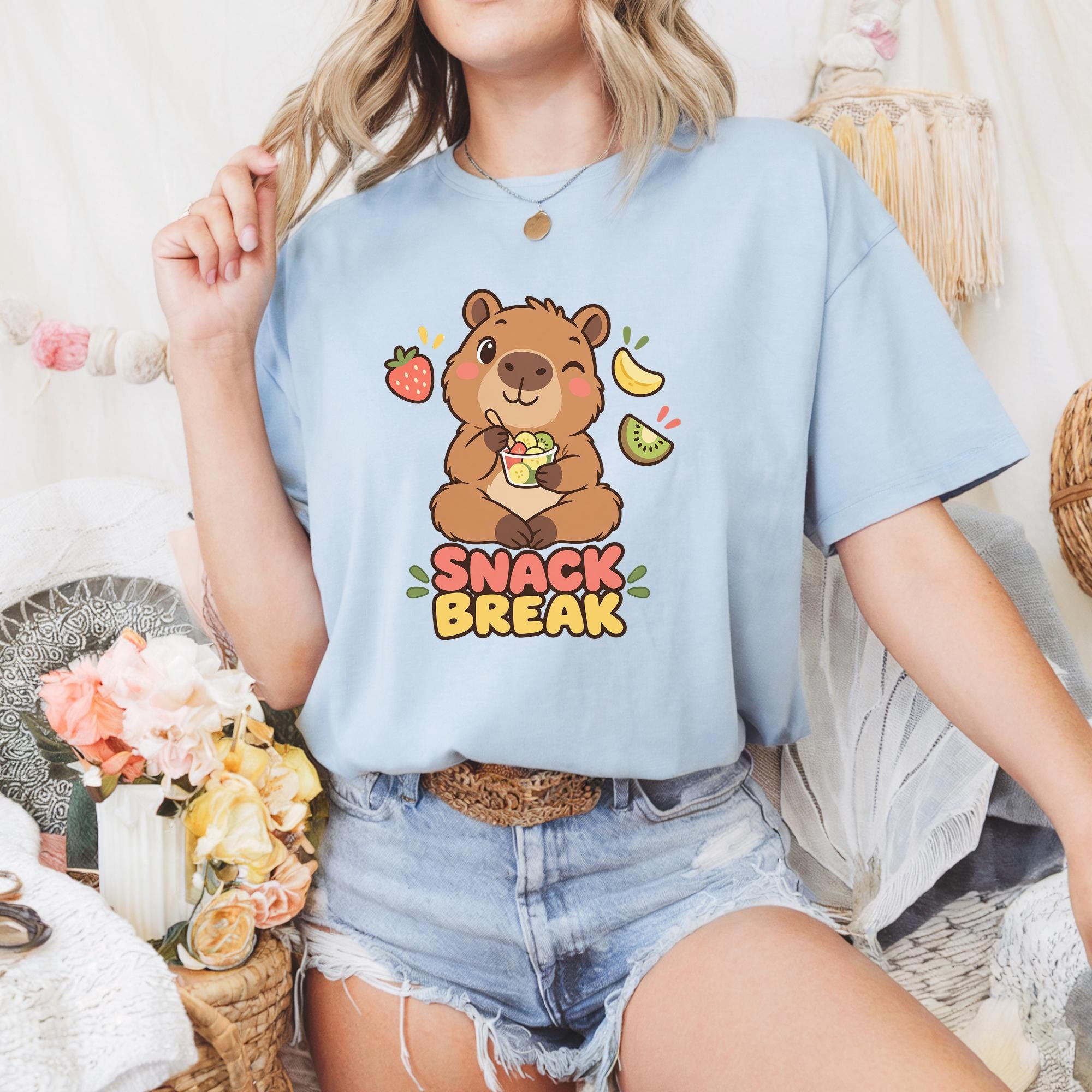 A woman wearing a blue t-shirt with a cartoon bear and the text "SNACK BREAK" is sitting in a room with a basket of flowers and a woven basket.