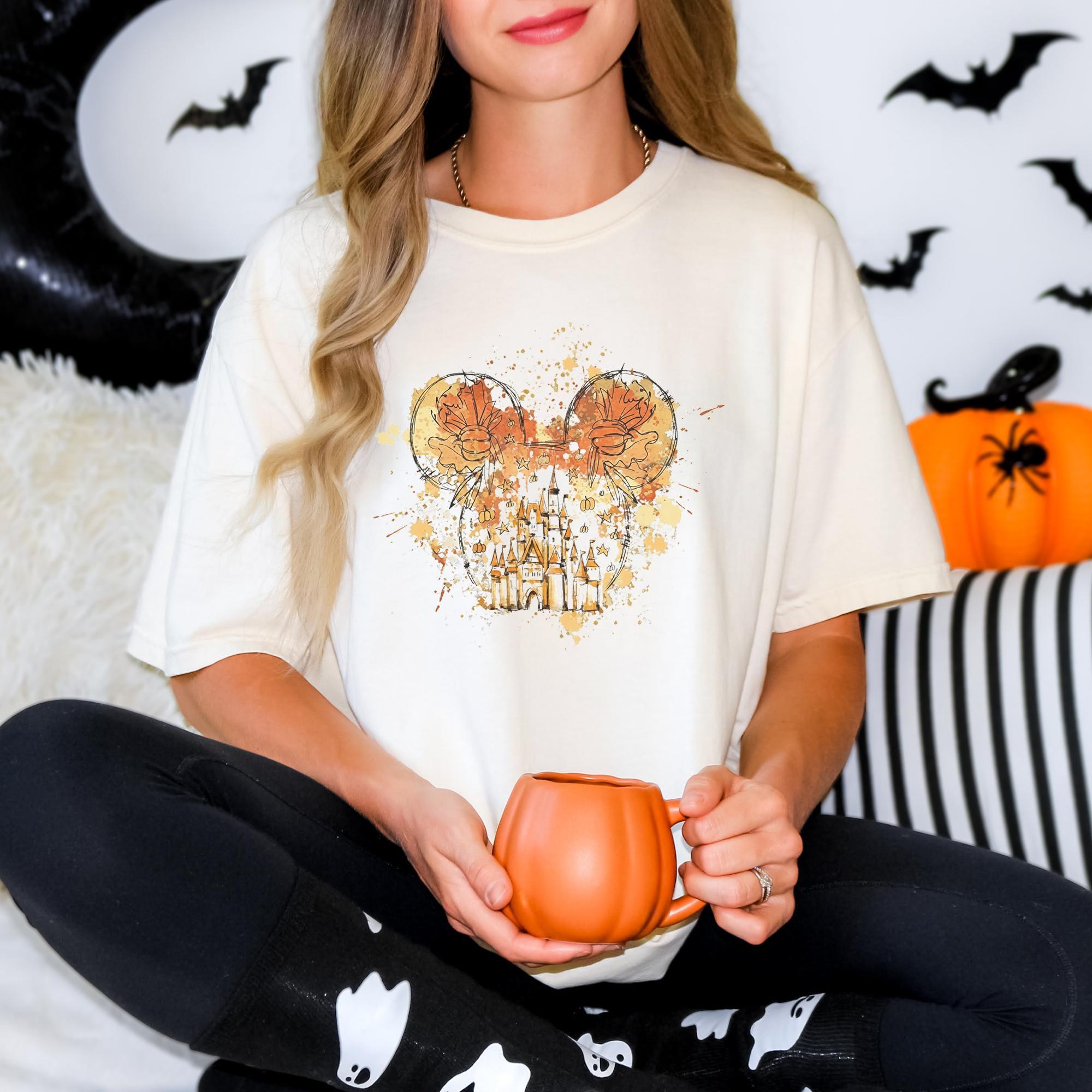 A woman wearing a white t-shirt with a graphic of Mickey Mouse ears holding a pumpkin-shaped mug, sitting on a couch with Halloween decorations in the background.