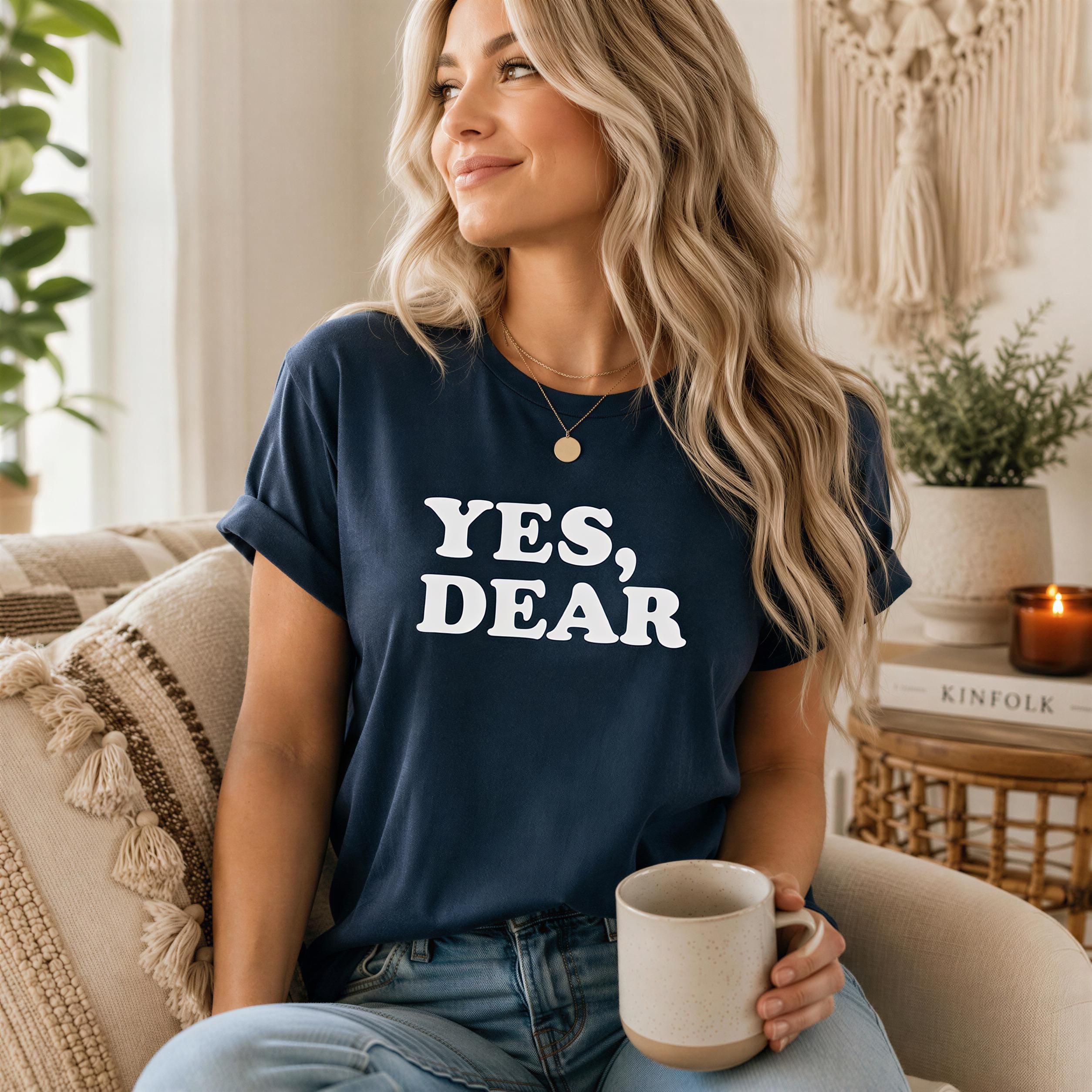A woman with long blonde hair is sitting on a couch, wearing a navy blue t-shirt with the text "YES, DEAR" printed on it. She is holding a mug in her hand.