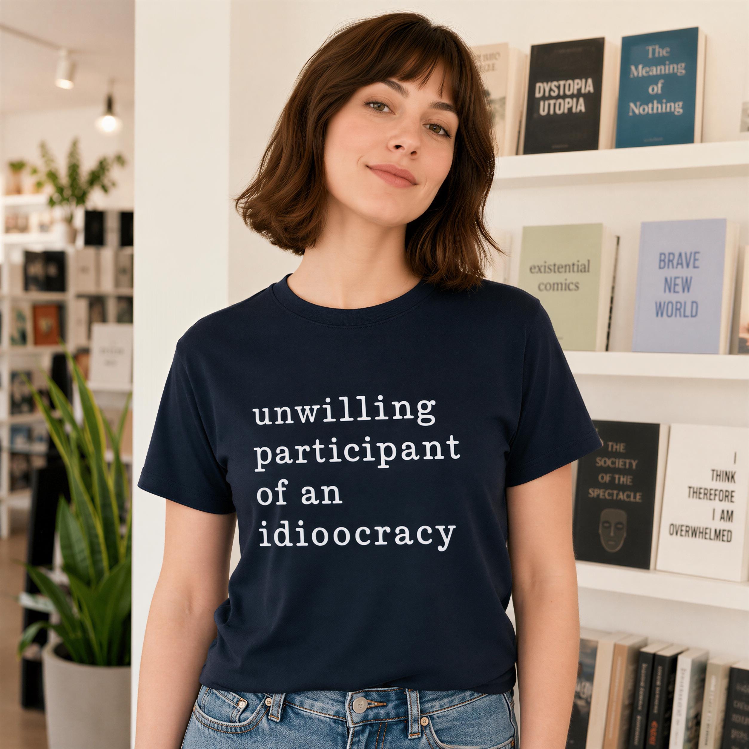 A woman wearing a navy blue t-shirt with the text "unwilling participant of an idioticocracy" stands in front of a bookshelf filled with various books.