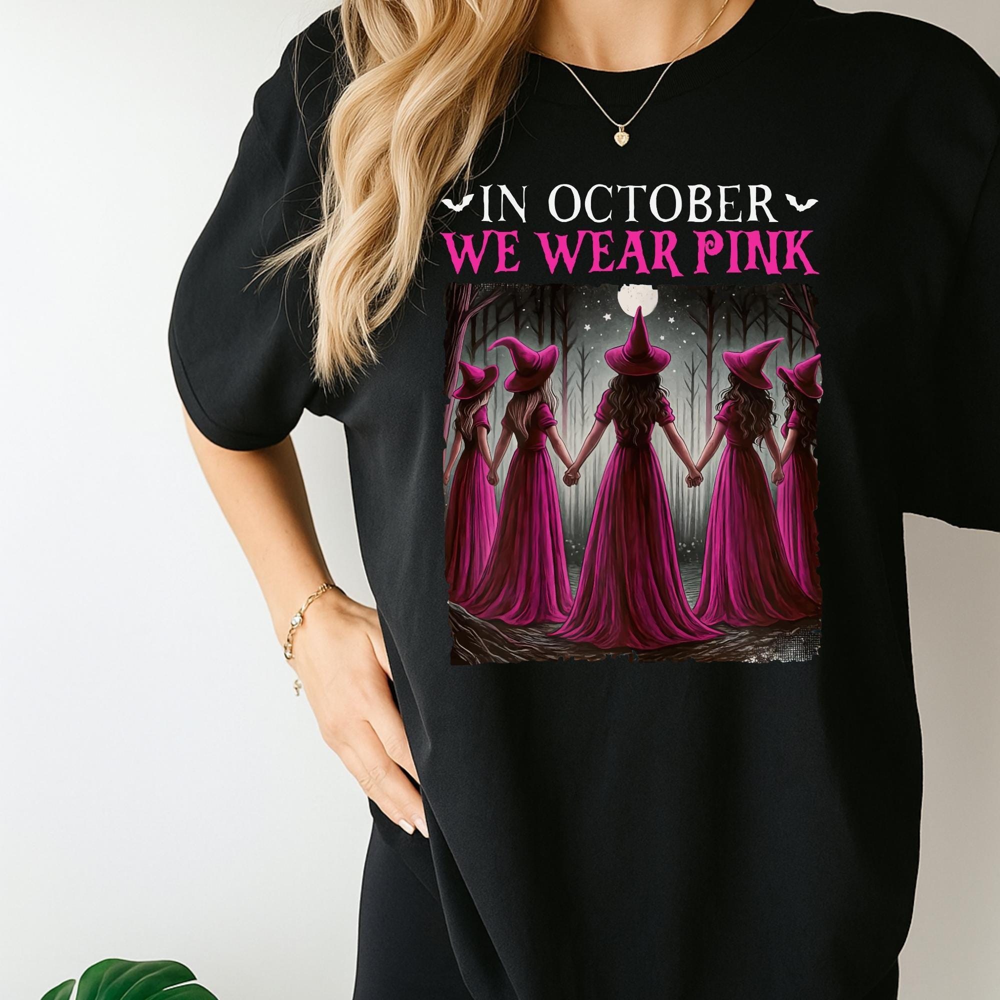 A woman wearing a black t-shirt with a graphic design featuring a group of women dressed in pink and witch hats, with the text "In October we wear pink" printed on the shirt.