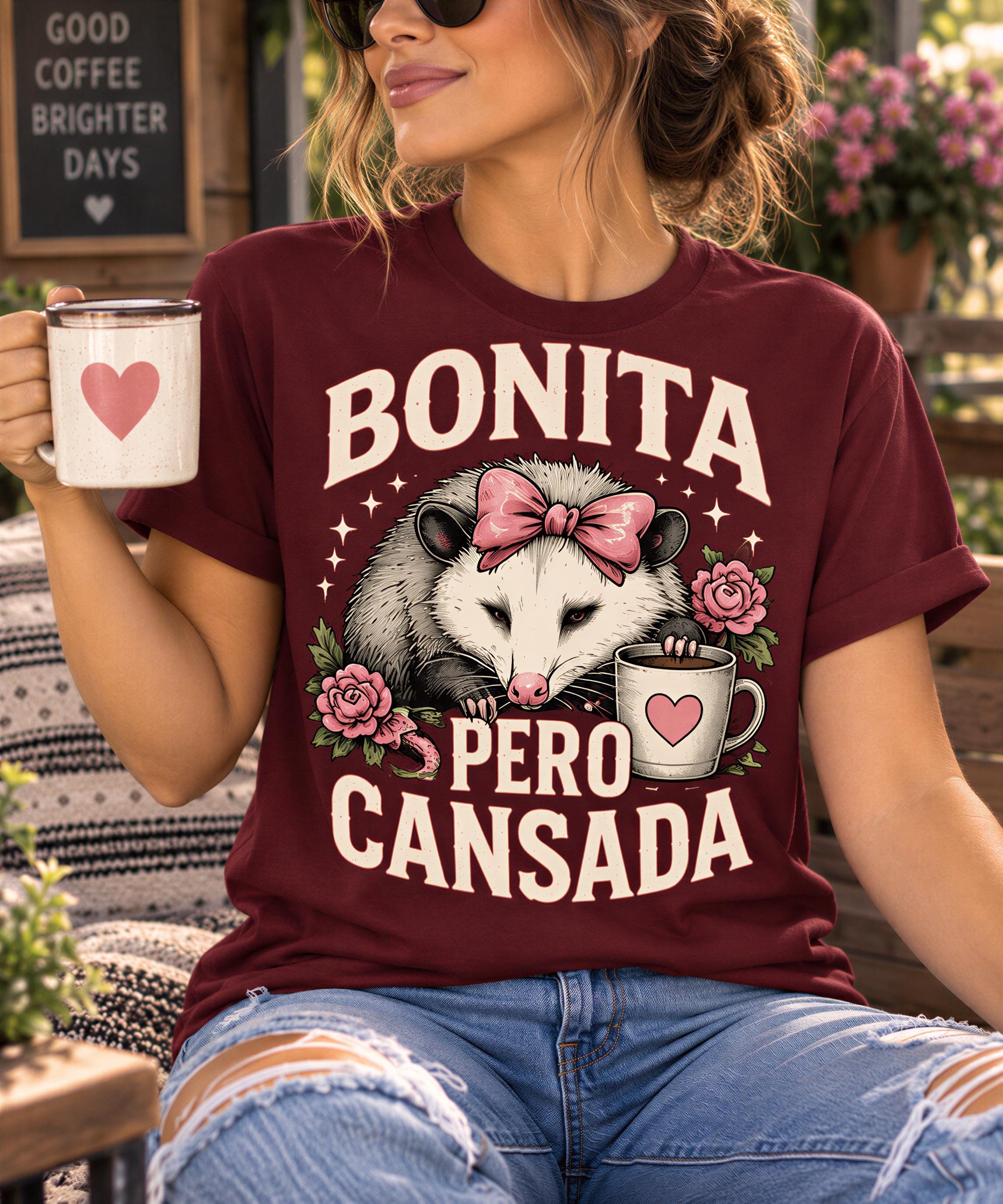A woman wearing a burgundy t-shirt with a graphic design of an opossum holding a coffee cup and flowers, along with the text "BONITA PERO CANSADA".