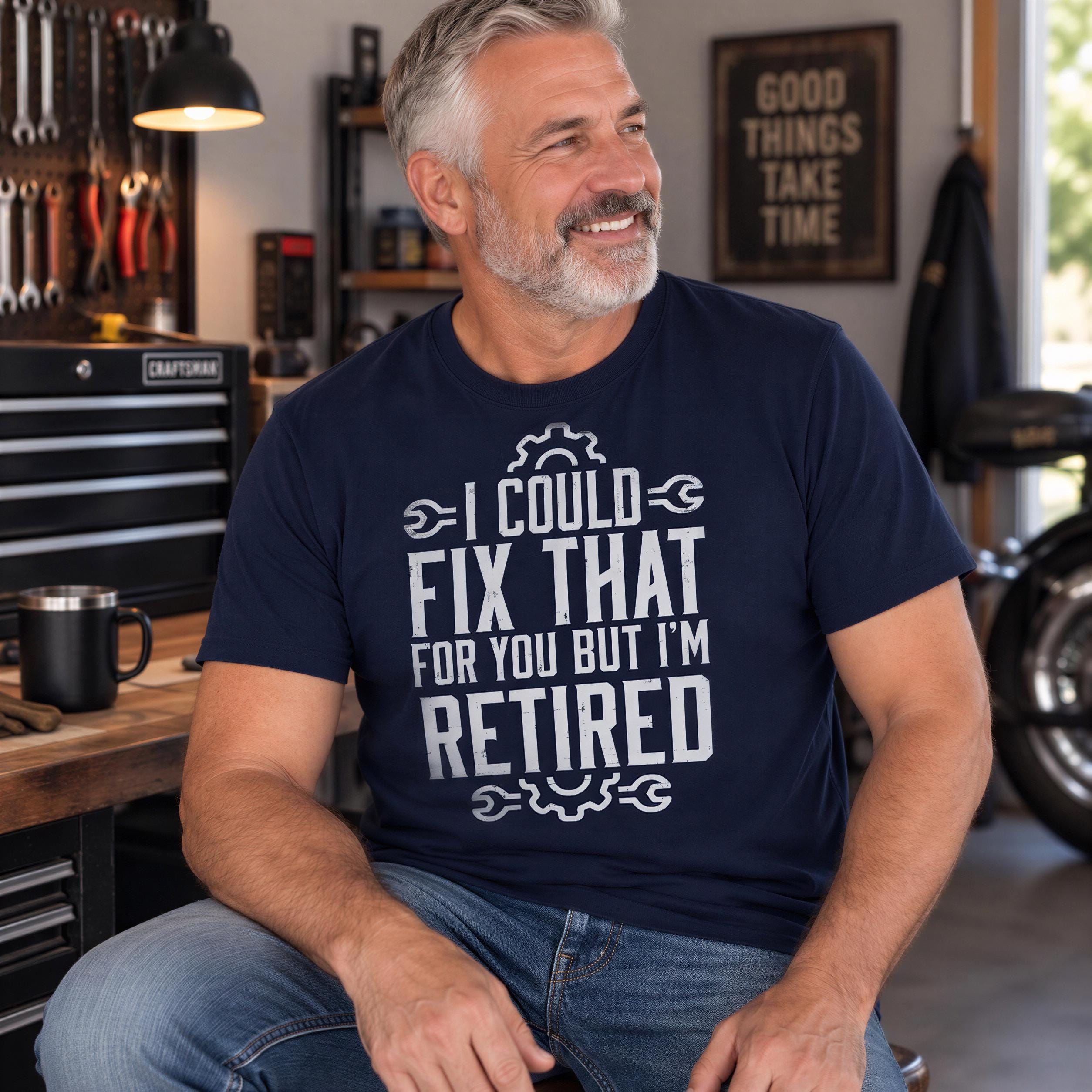 A man wearing a navy blue t-shirt with a humorous text printed on it, sitting in a workshop with various tools and equipment visible in the background.