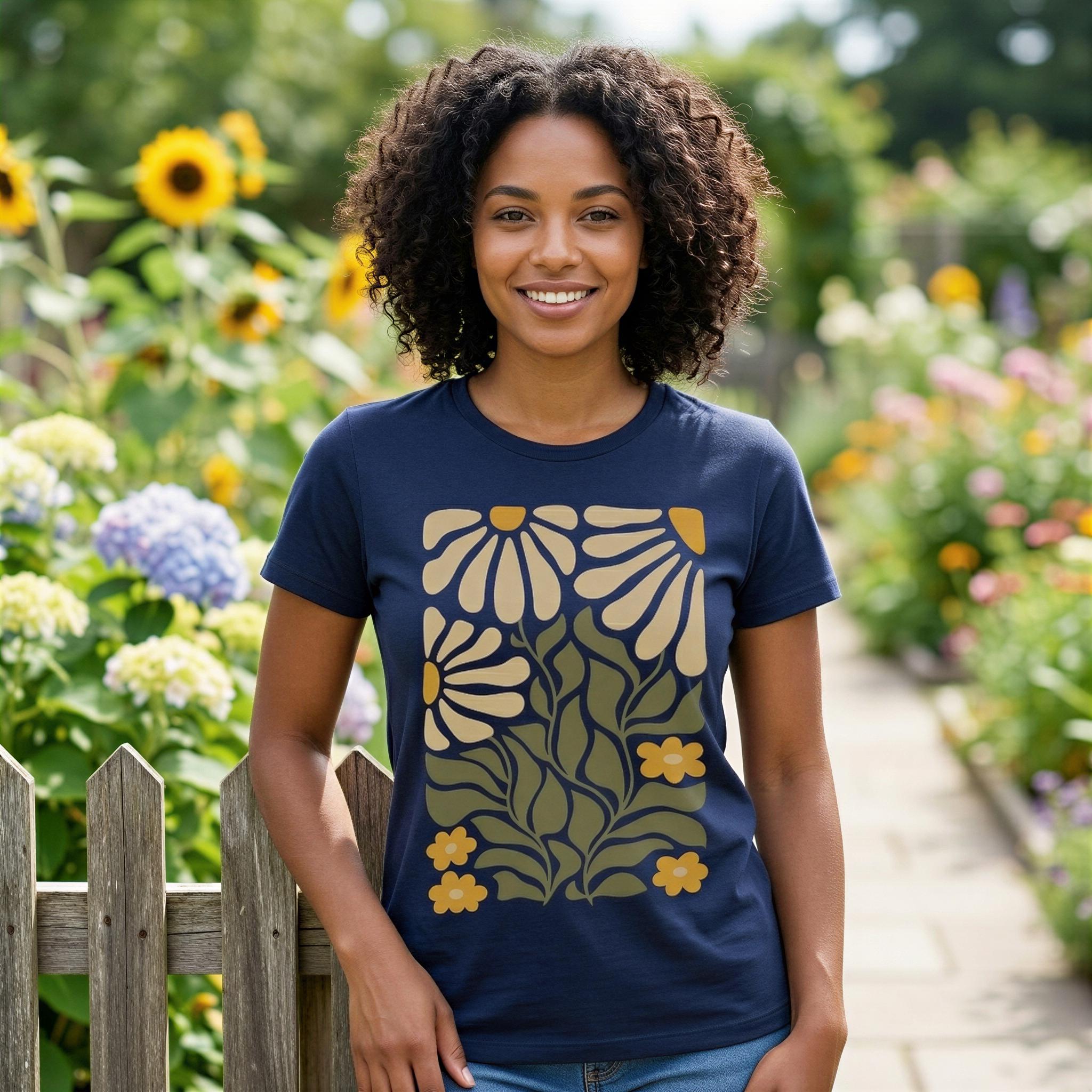 A smiling woman with curly hair stands in a garden surrounded by colorful flowers, wearing a navy blue t-shirt with a floral design.