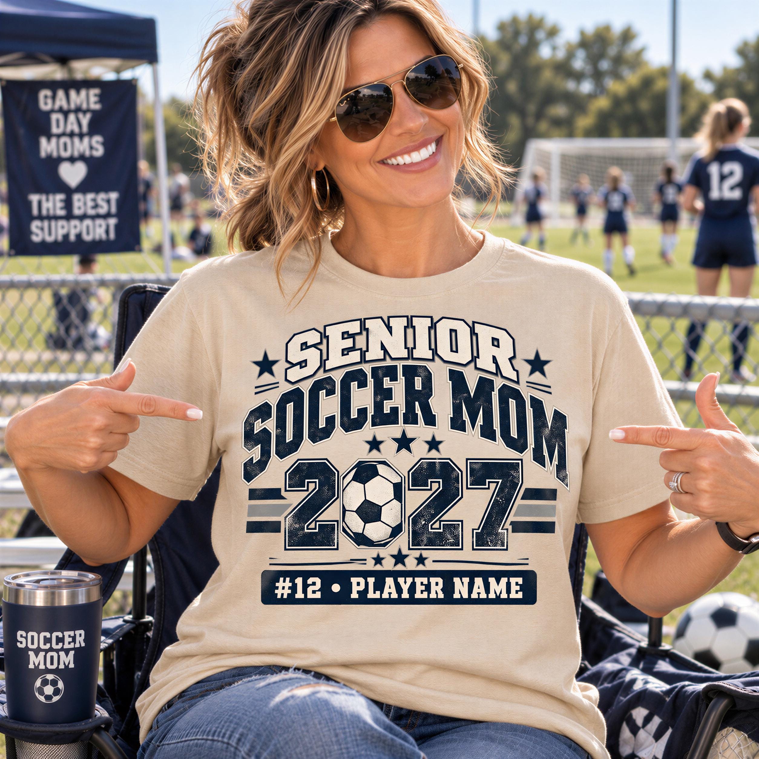 A smiling woman wearing a t-shirt with a soccer theme and pointing at her shirt, sitting in a chair at a sports event.
