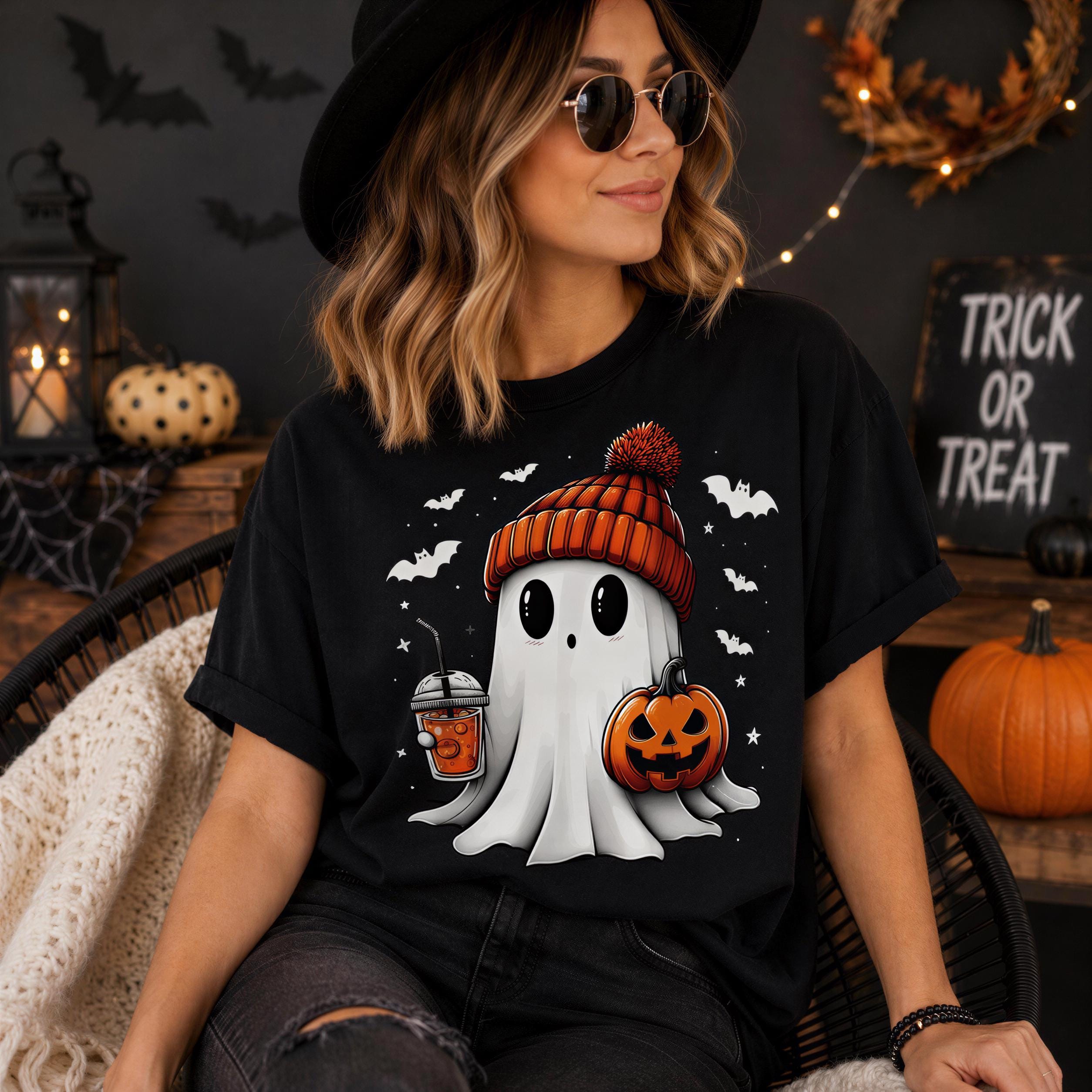 A woman wearing a black hat and sunglasses is sitting in a chair, with a Halloween-themed t-shirt featuring a ghost, pumpkin, and a drink.