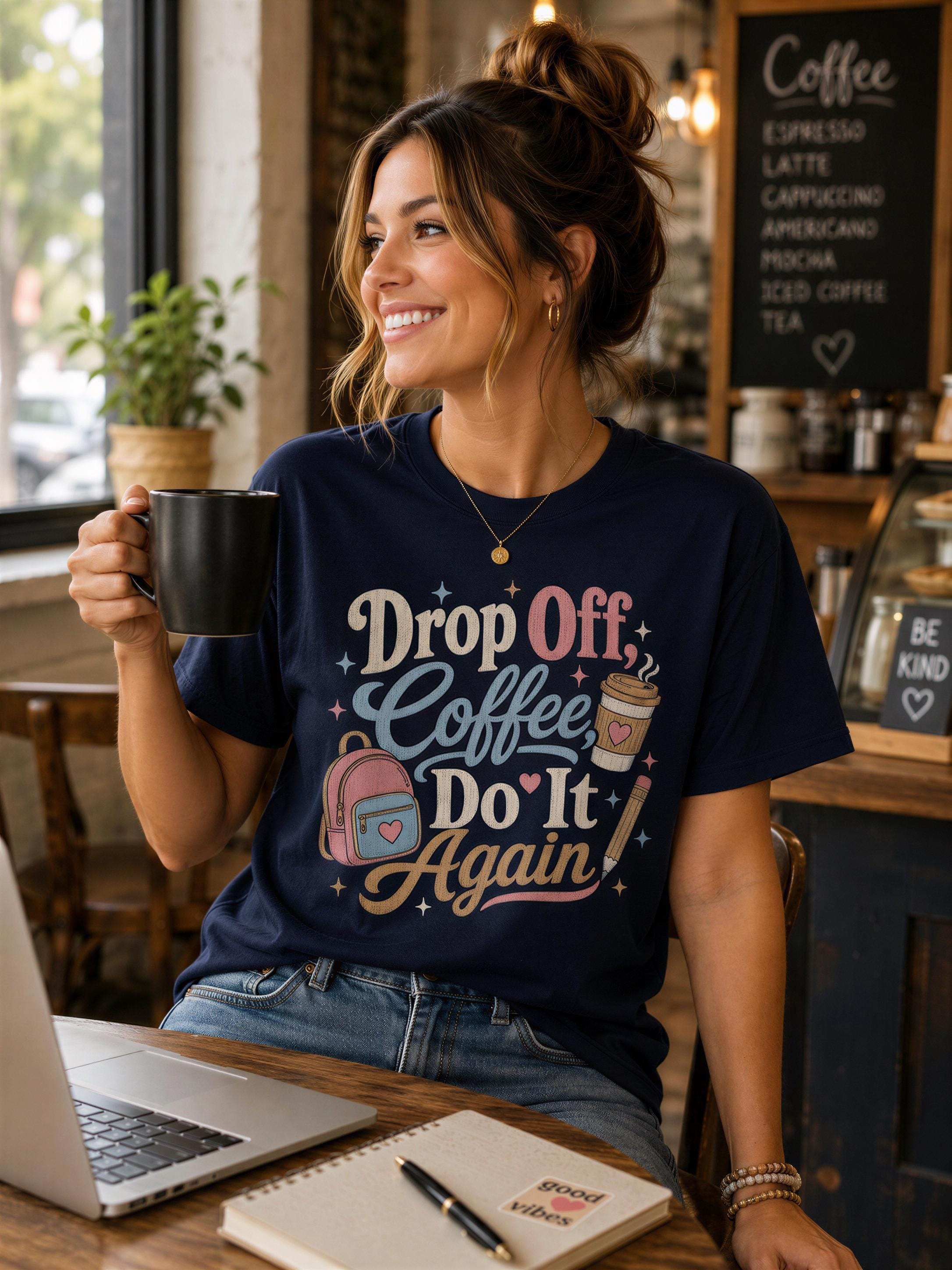 A woman wearing a blue t-shirt with a colorful design is sitting at a table in a coffee shop, holding a black coffee mug and smiling.