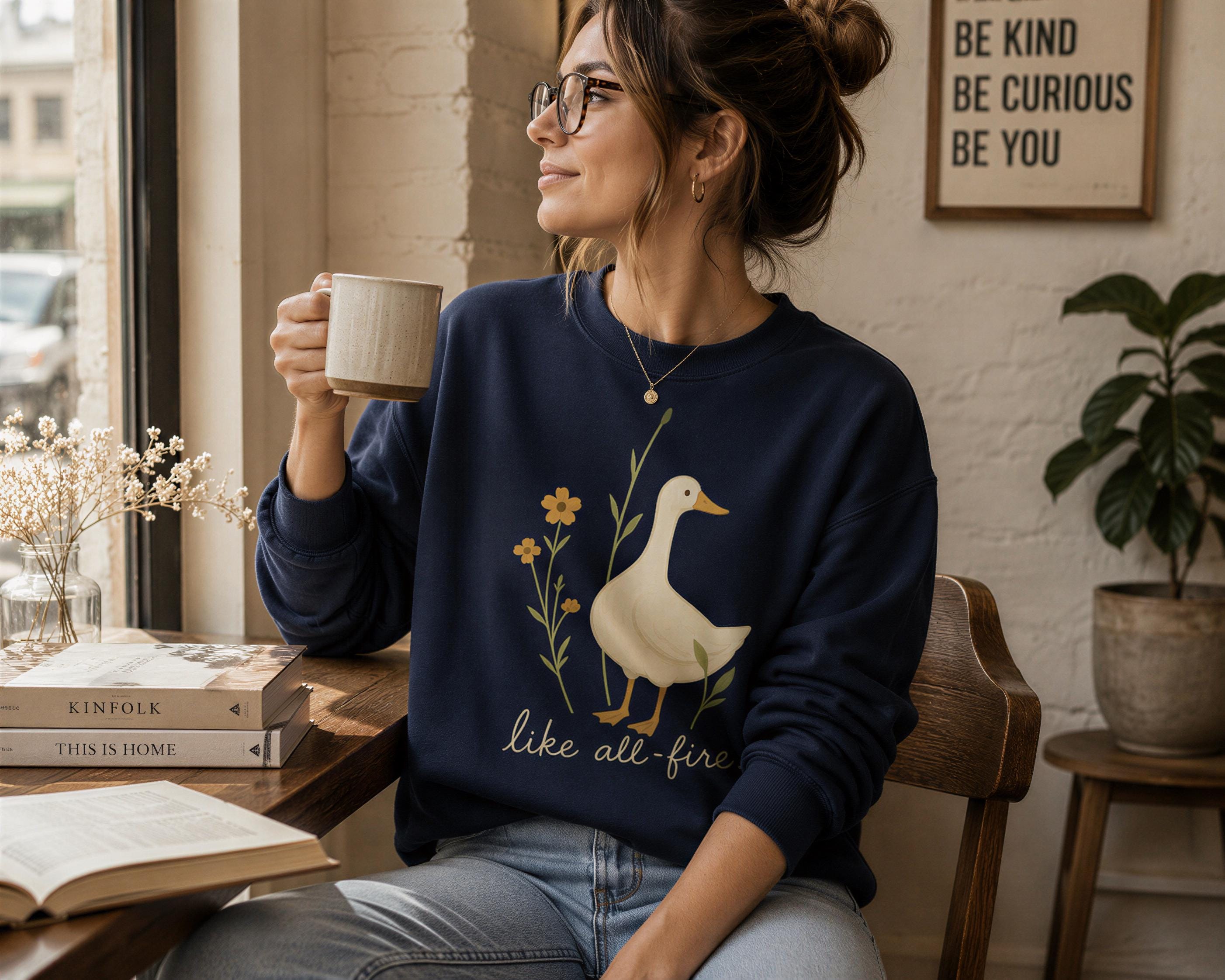 A woman wearing a blue sweatshirt with a duck design and the text "like all fire" is sitting at a table, holding a coffee mug and reading a book.