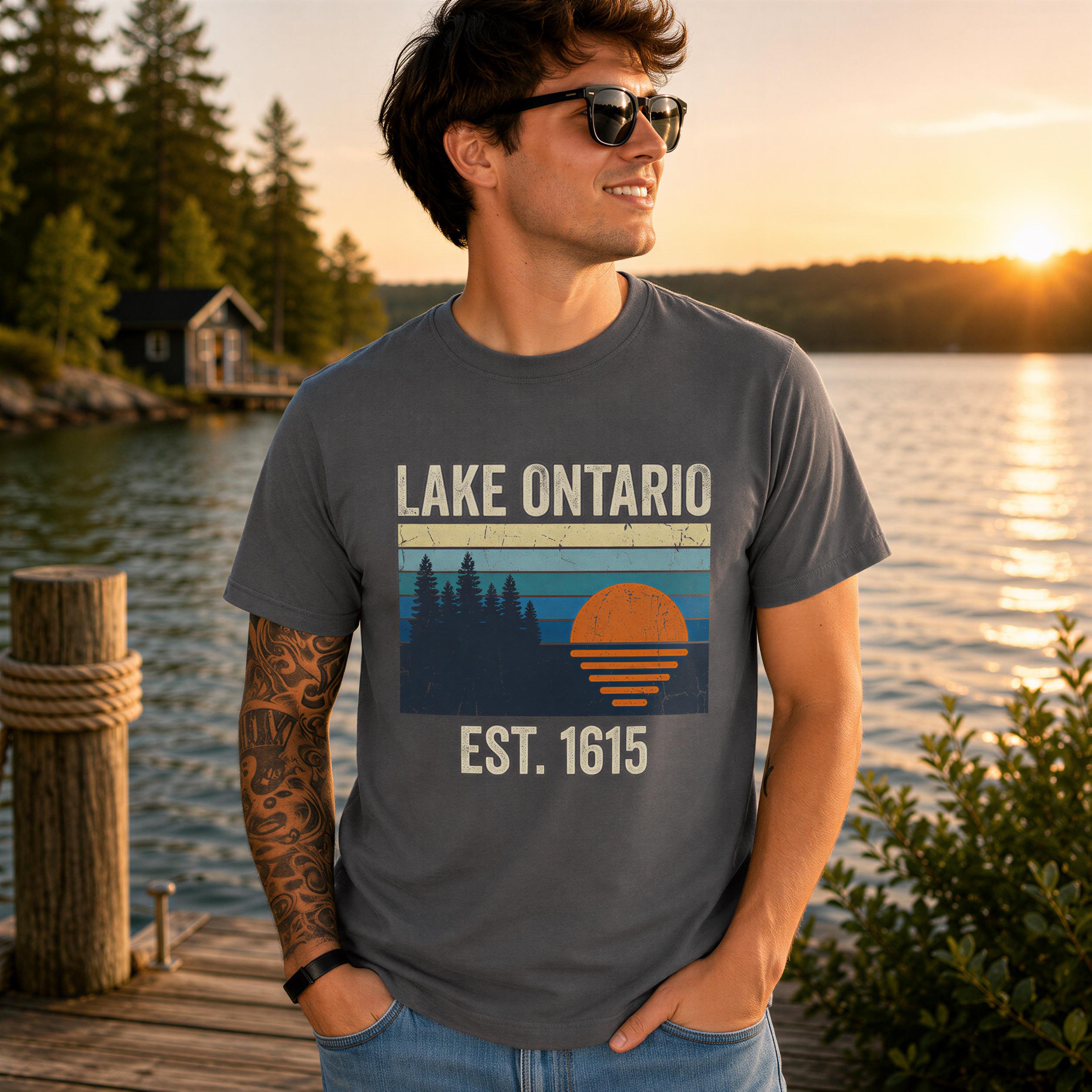 A man wearing a gray t-shirt with a graphic design of a sunset and trees, standing on a dock overlooking a lake.