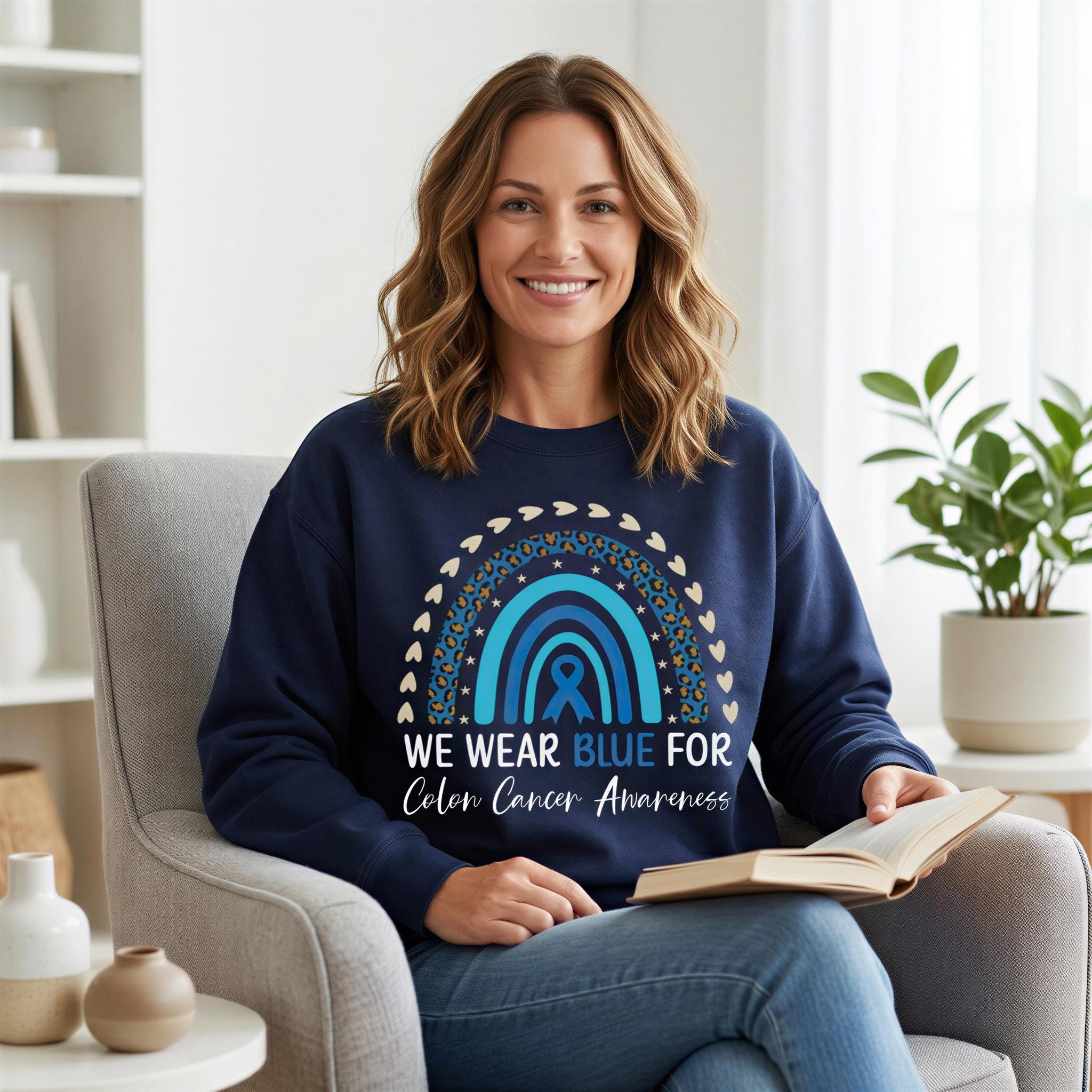 A woman is sitting in a chair, smiling and holding an open book. She is wearing a navy blue sweatshirt with a rainbow design and the text "WE WEAR BLUE FOR COLON CANCER AWARENESS".