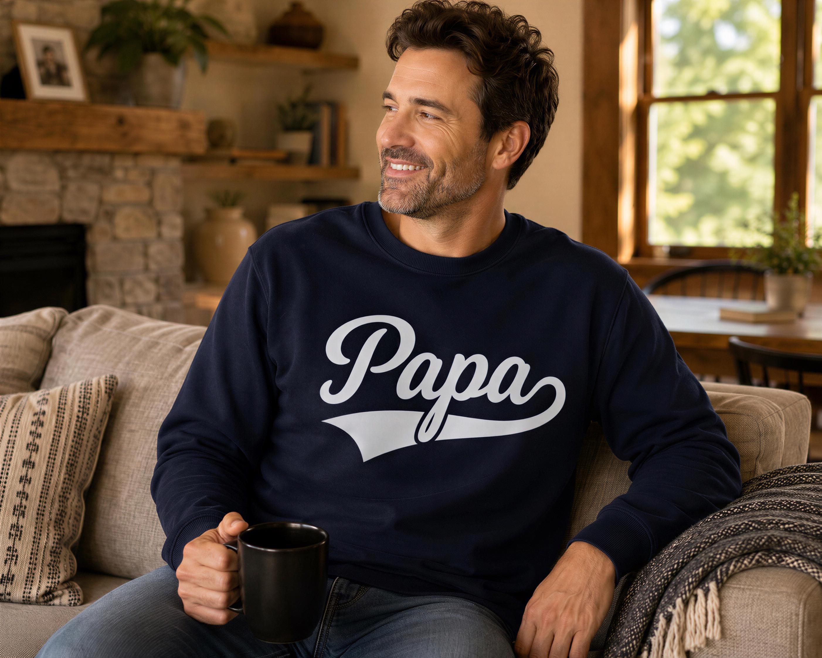 A man wearing a navy blue sweatshirt with the word "Papa" printed on it is sitting on a couch, holding a black mug and smiling.