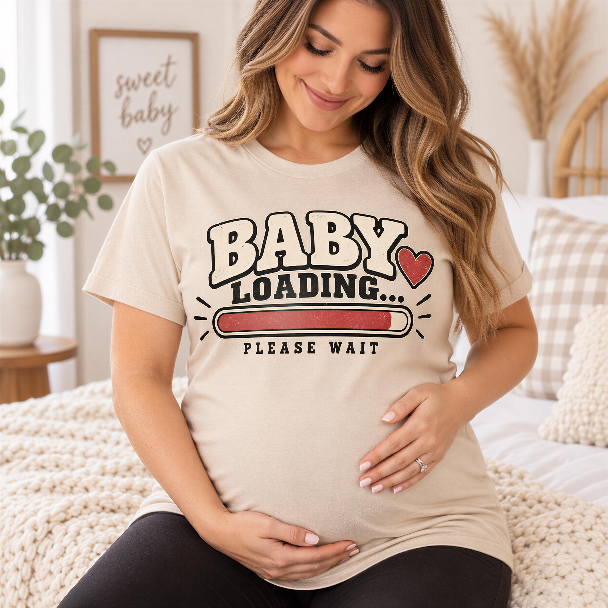 A pregnant woman wearing a t-shirt with the text "BABY LOADING... PLEASE WAIT" is sitting on a bed.