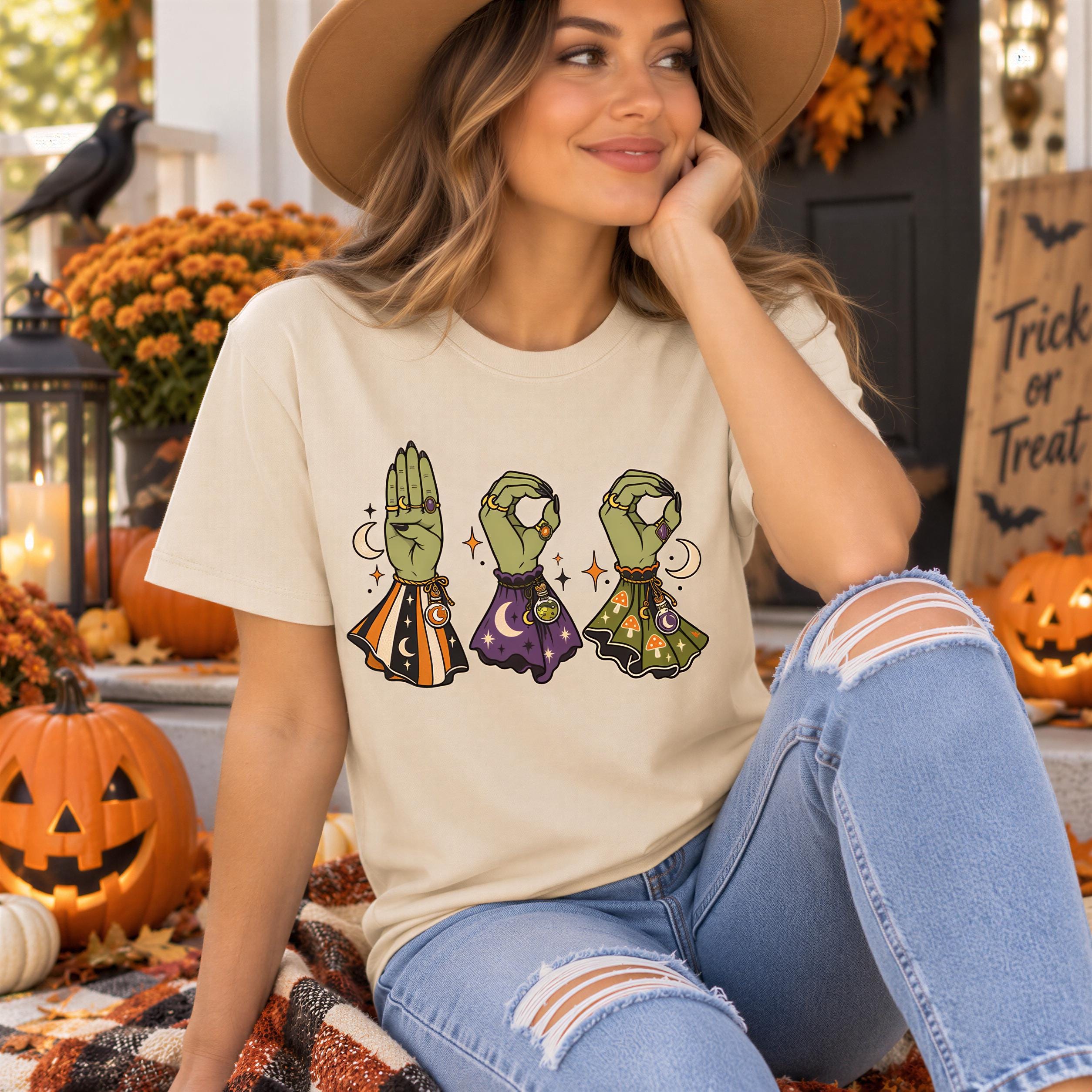 A woman wearing a white t-shirt with a graphic design of three green alien hands holding a crystal ball, surrounded by Halloween decorations such as pumpkins and bats.