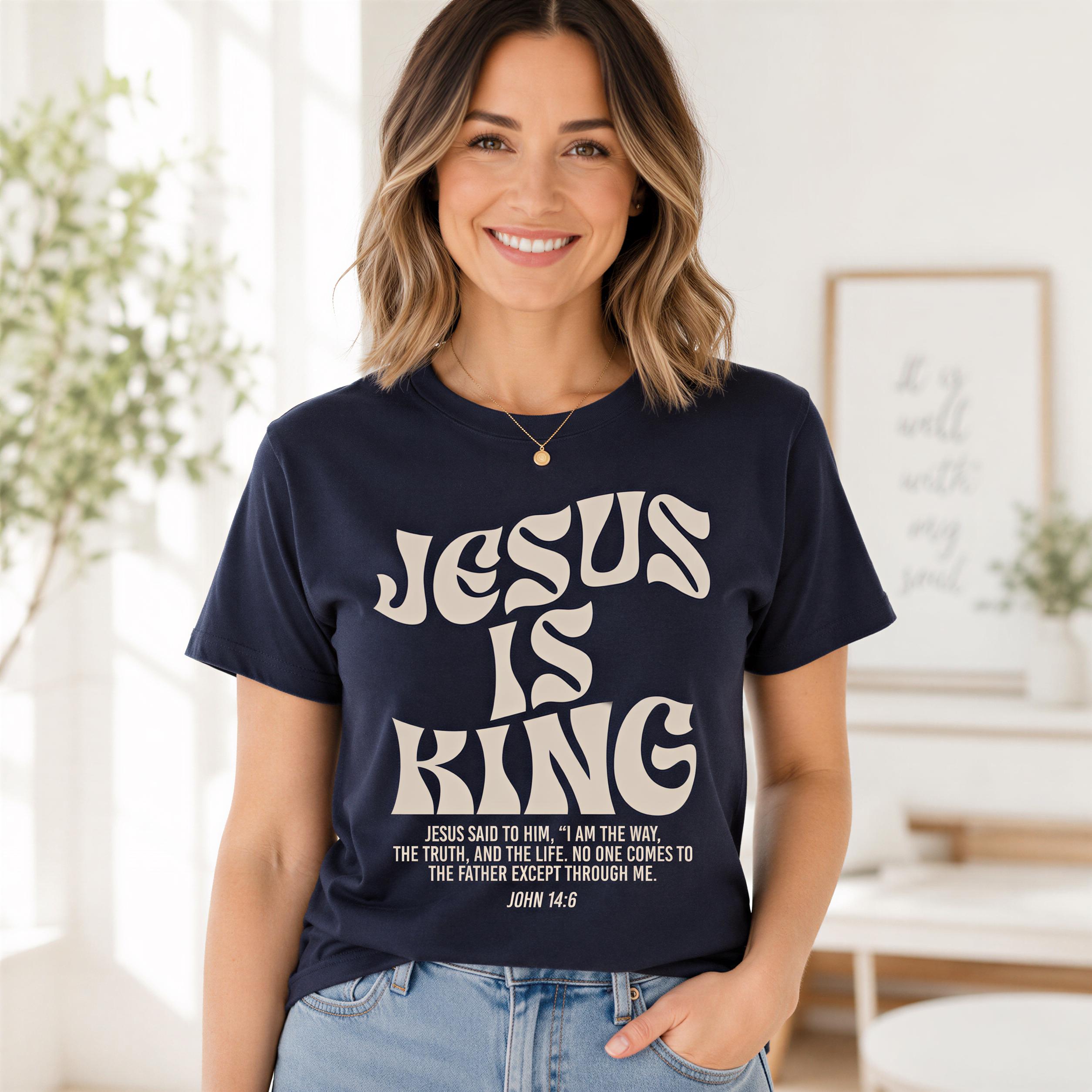 A woman wearing a navy blue t-shirt with the text "JESUS IS KING" and a Bible verse from John 14:6, smiling at the camera.