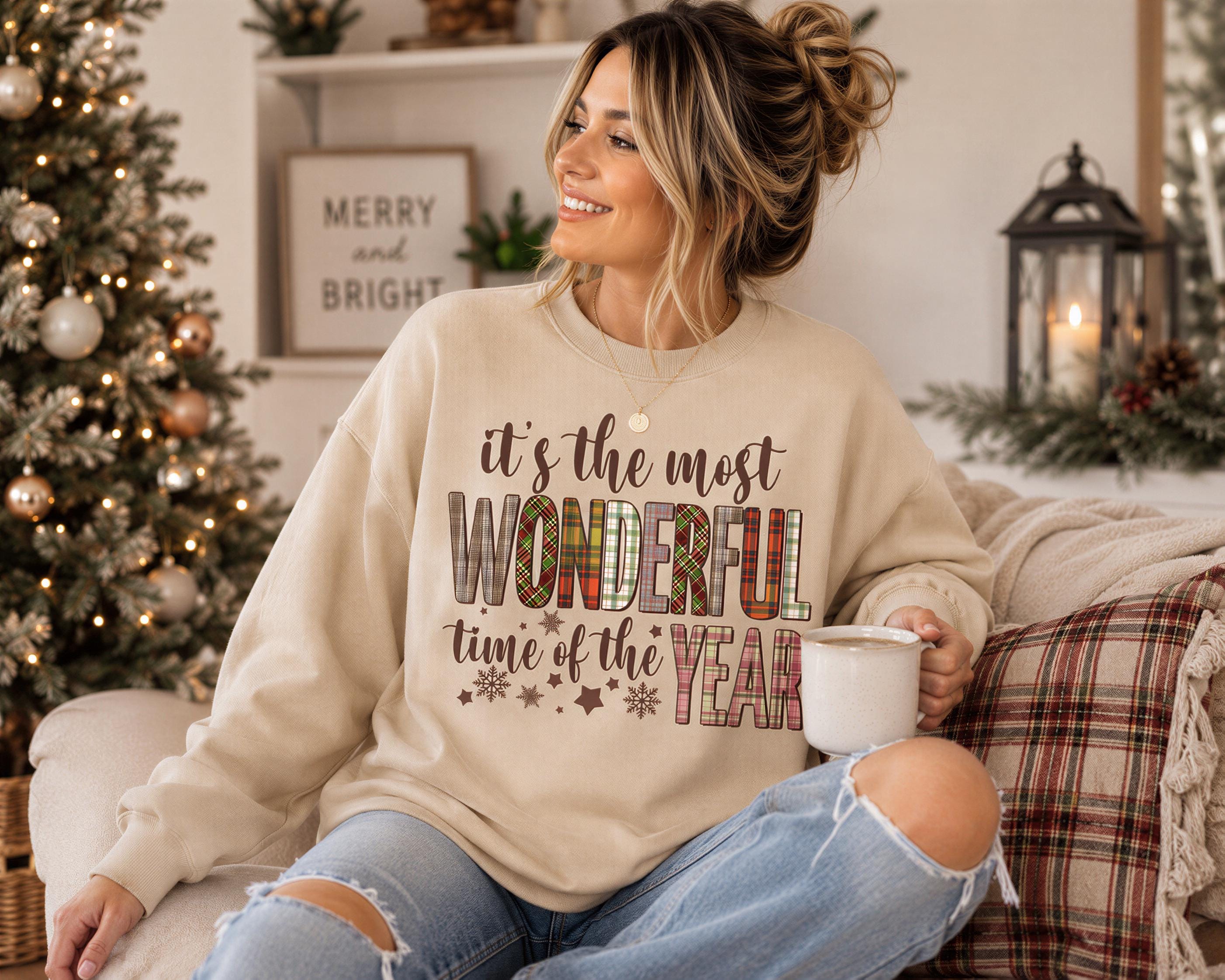 A woman sitting on a couch, wearing a beige sweatshirt with a Christmas-themed design, smiling and holding a mug.
