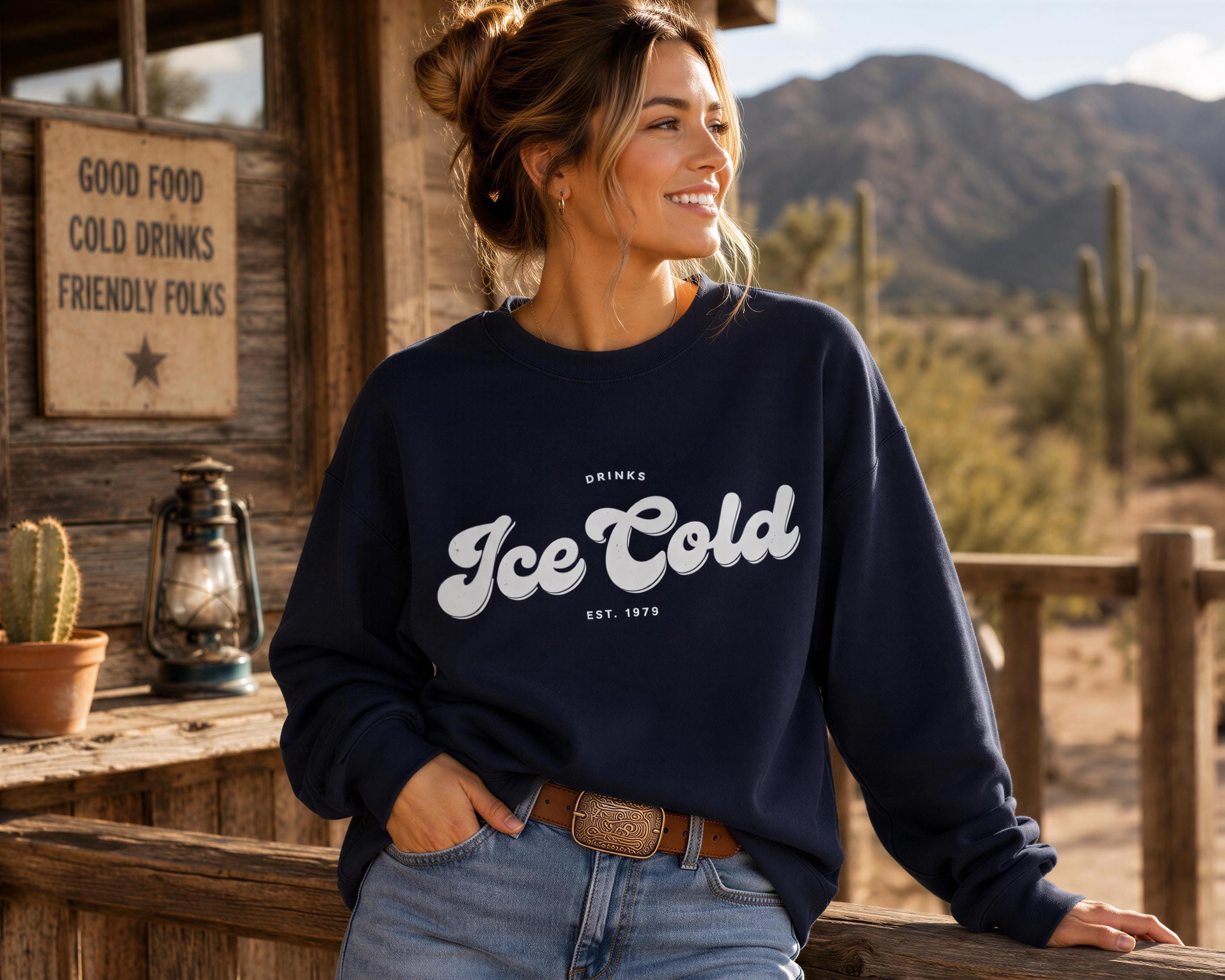 A woman wearing a navy blue sweatshirt with the text "Drink Ice Cold" stands in front of a wooden building with a sign that says "Good Food Cold Drinks Friendly Folks".