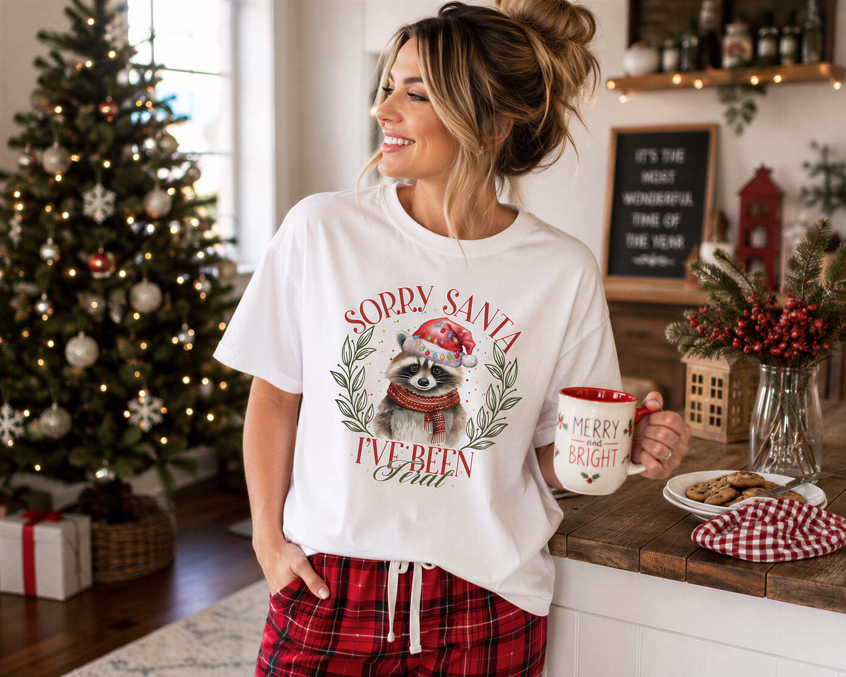 A woman wearing a white t-shirt with a graphic of a raccoon and the text "Sorry Santa, I've been bad" is standing in a kitchen, holding a mug and smiling.