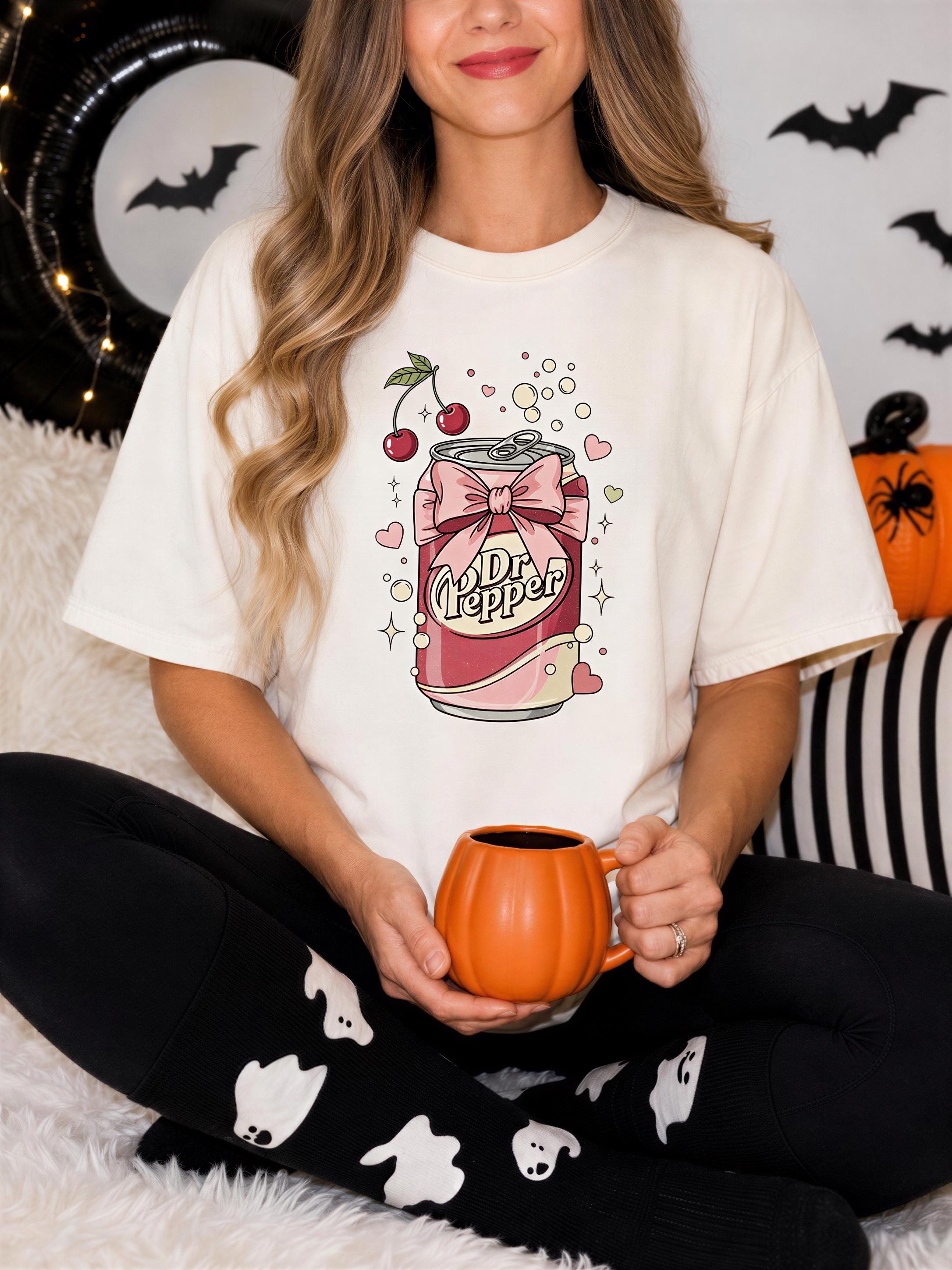 A woman wearing a white t-shirt with a graphic of a can of Dr Pepper and a bow, holding an orange mug, sitting on a bed with Halloween decorations in the background.