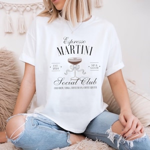 Peut inclure: T-shirt blanc avec l'inscription "Espresso Martini Social Club" et un dessin de cocktail. Le design comprend les mots "EST 2024" et "Sip & Savor". Le t-shirt est porté avec un jean bleu.