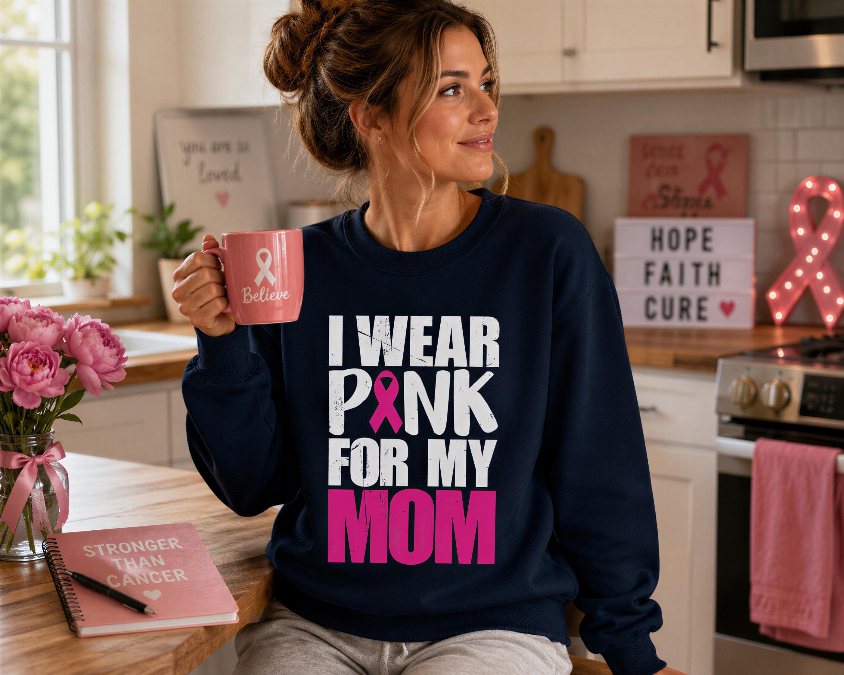 A woman wearing a navy blue sweatshirt with pink text and a pink ribbon, holding a pink mug, and sitting in a kitchen with a pink ribbon sign and flowers in the background.