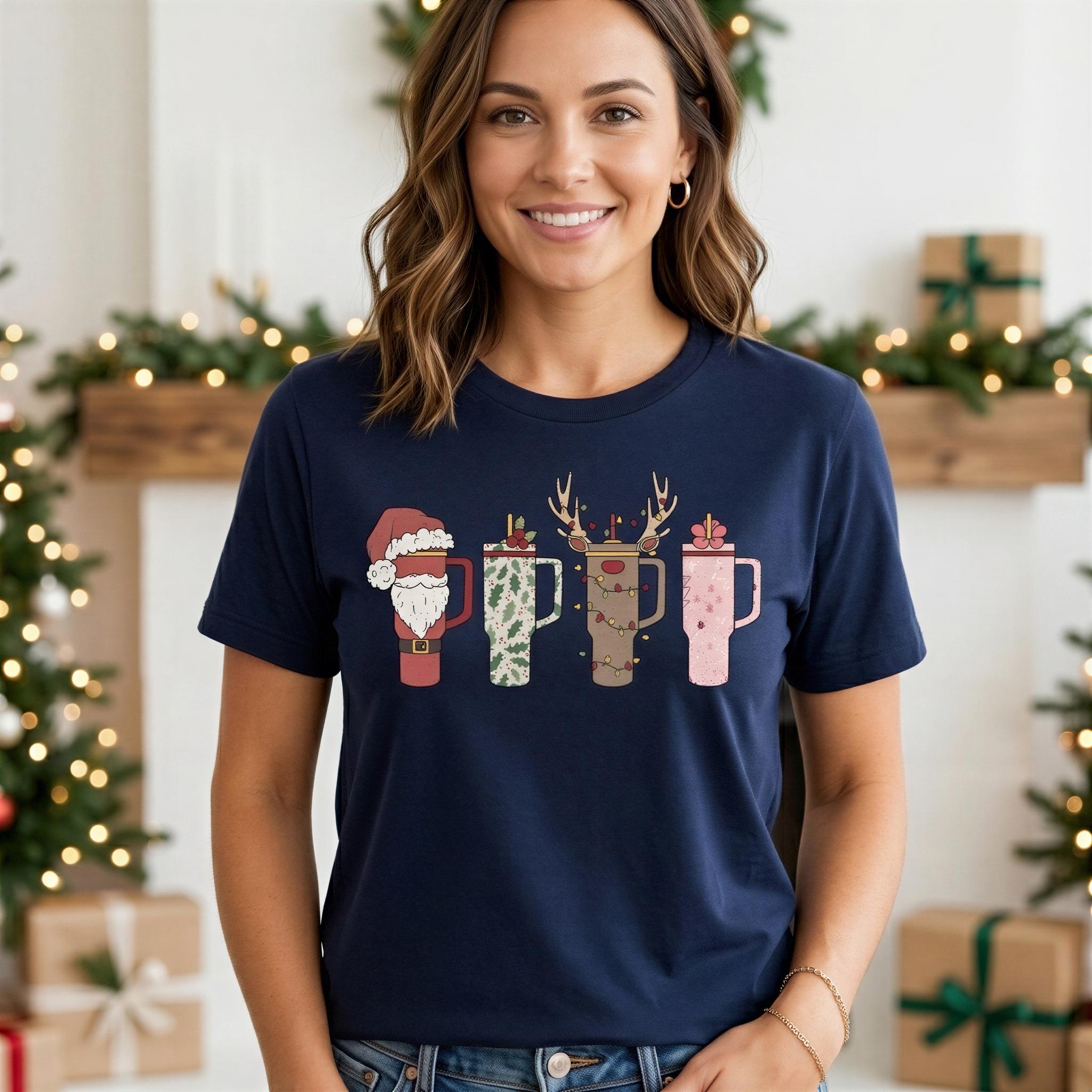 A woman wearing a navy blue t-shirt with a graphic design of four cups with Santa Claus hats on them, standing in front of a Christmas tree and gift boxes.