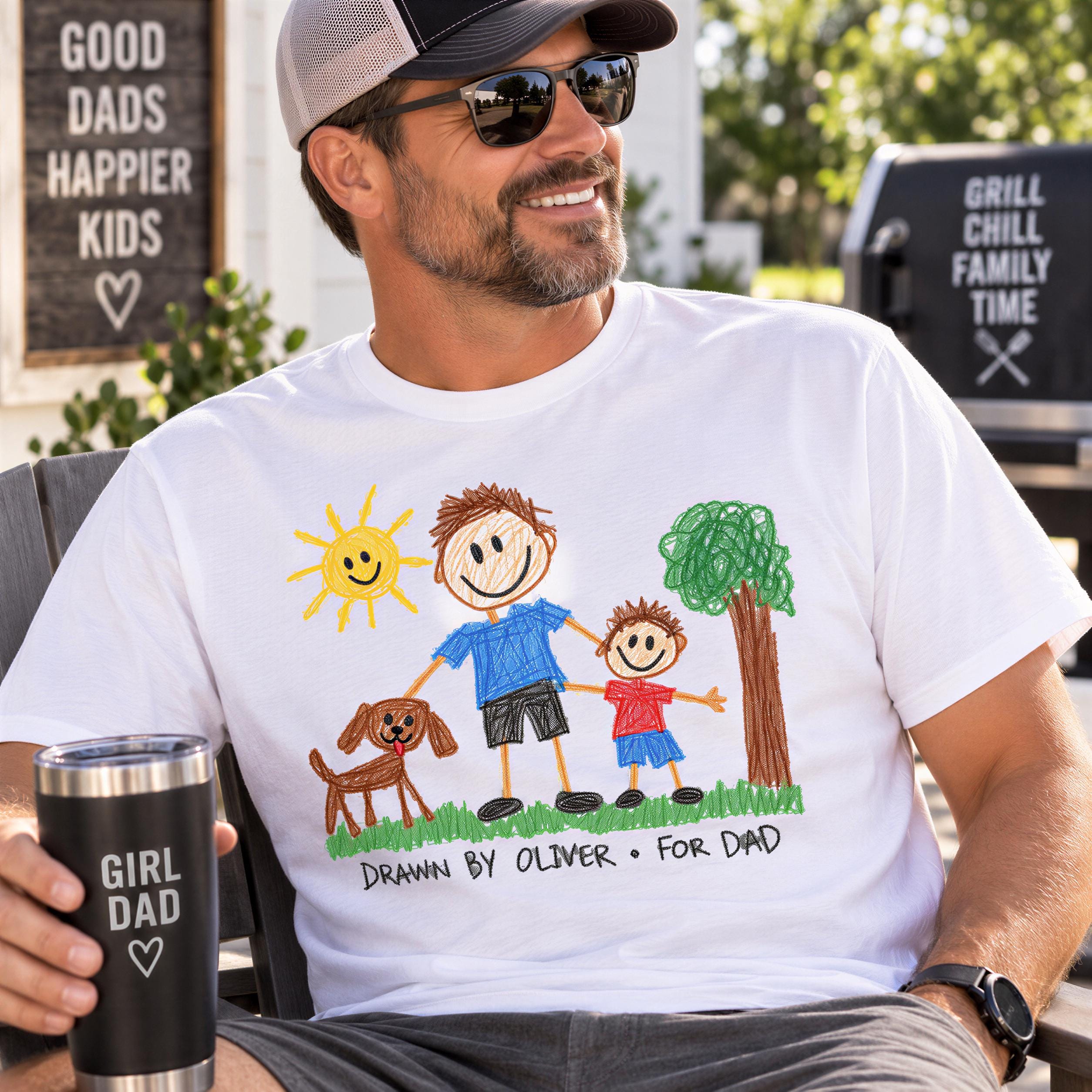 A man wearing a white t-shirt with a drawing of a father and son, sitting outdoors and holding a cup with a message that says "GIRL DAD DRAWN BY OLIVER FOR DAD".