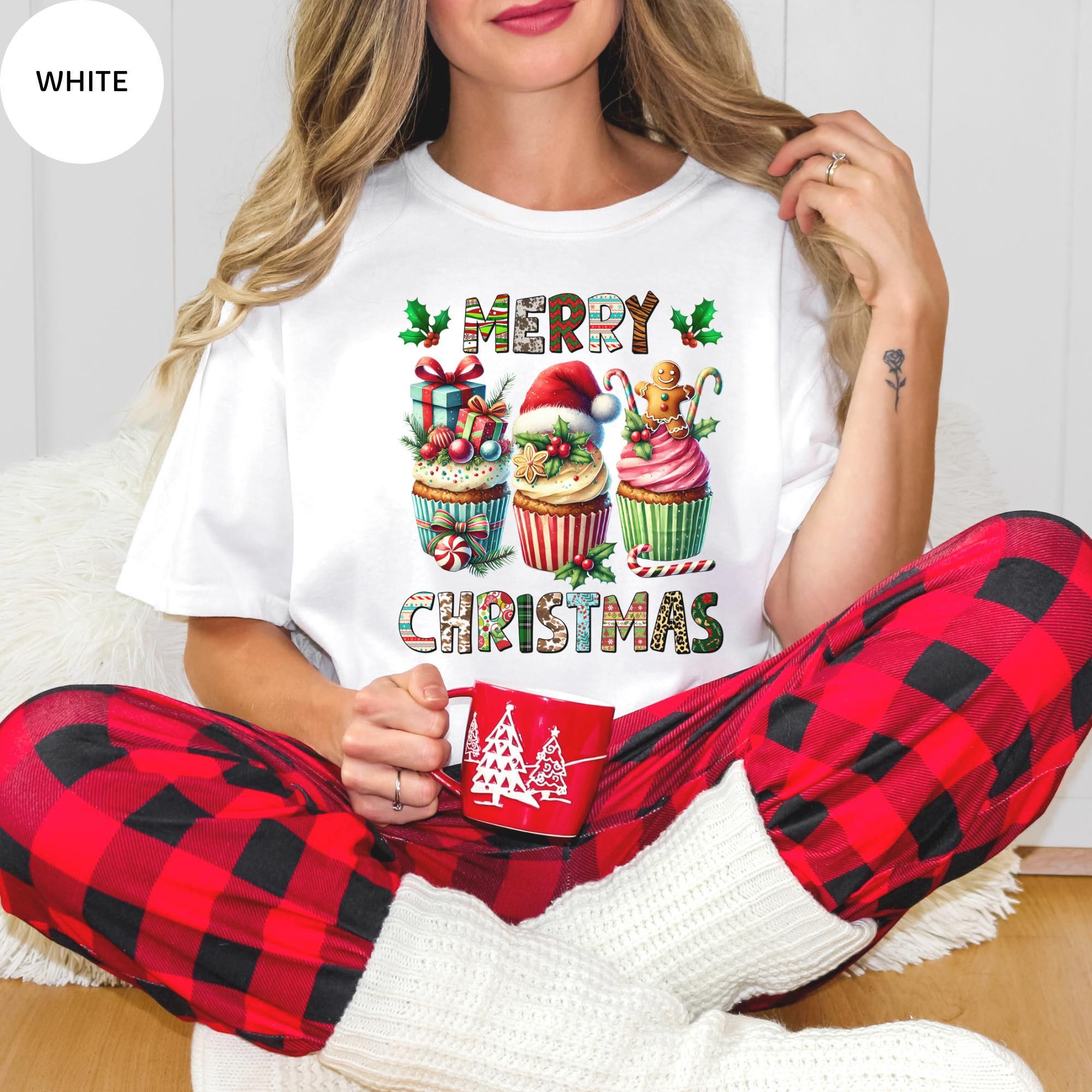 A woman wearing a white t-shirt with a Christmas-themed graphic and holding a red mug, sitting on the floor.