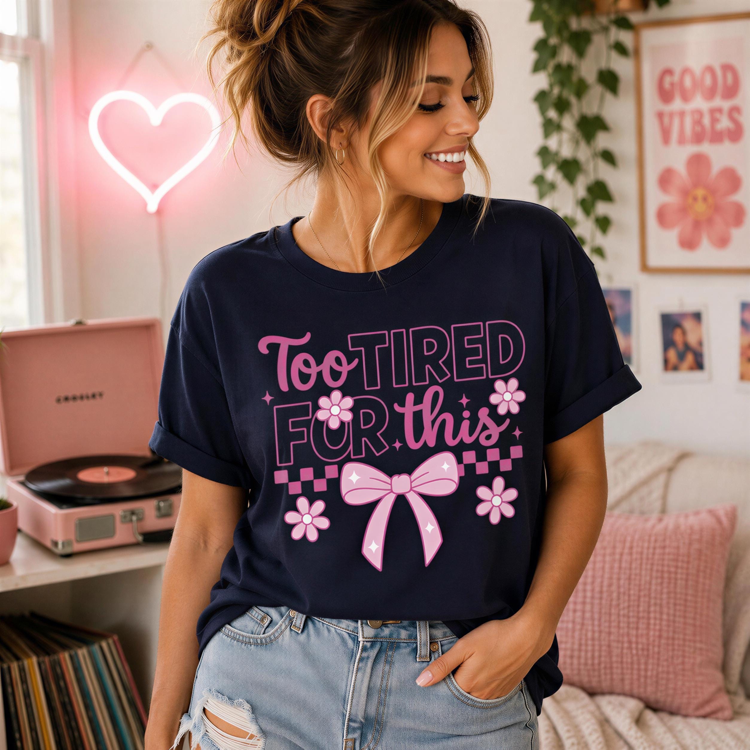 A woman wearing a black t-shirt with a pink bow and text, smiling and posing in a room with a pink record player and other decor.