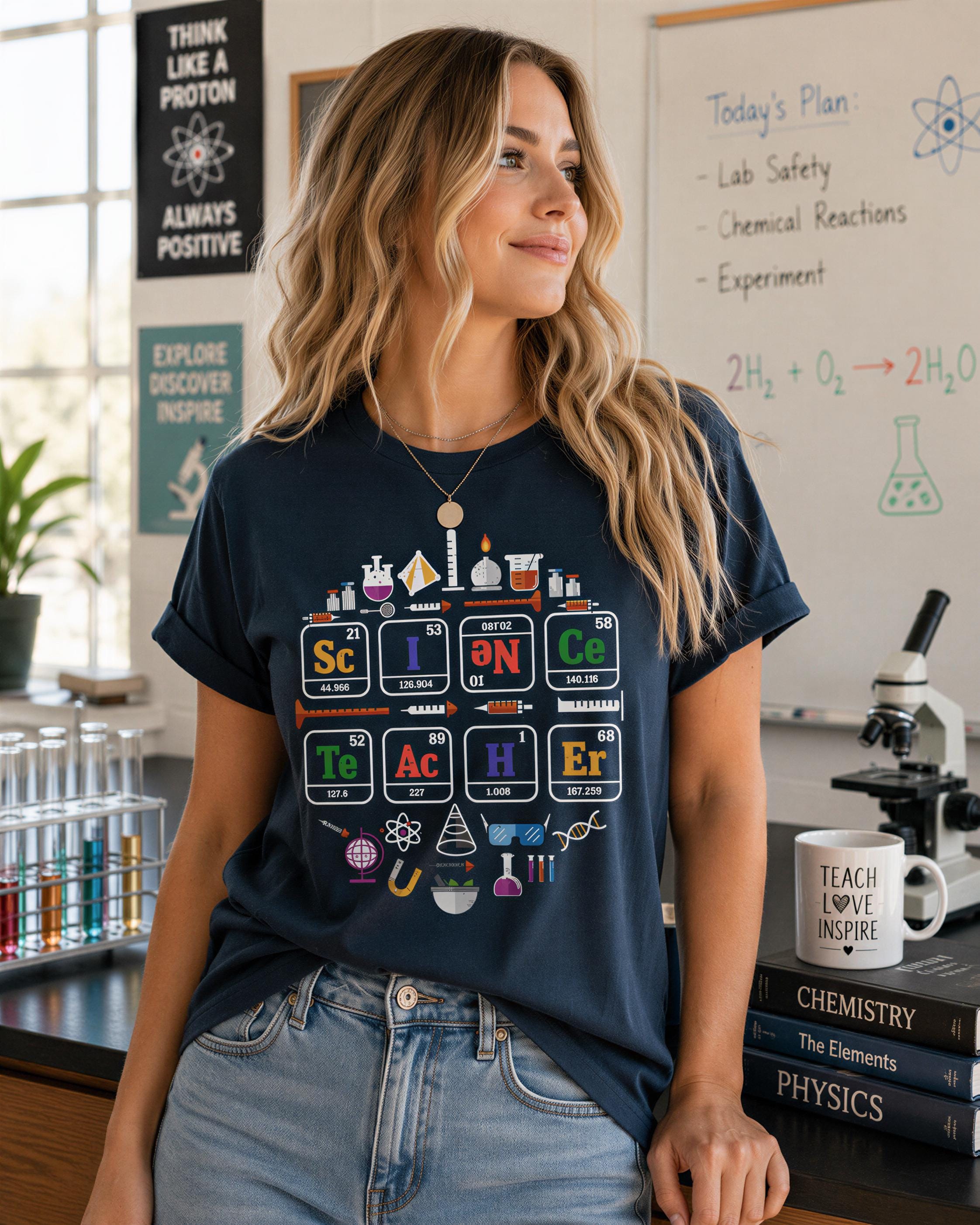 A woman wearing a blue t-shirt with a graphic design of the periodic table and various science-related icons, standing in a classroom setting with a whiteboard and laboratory equipment visible in the background.