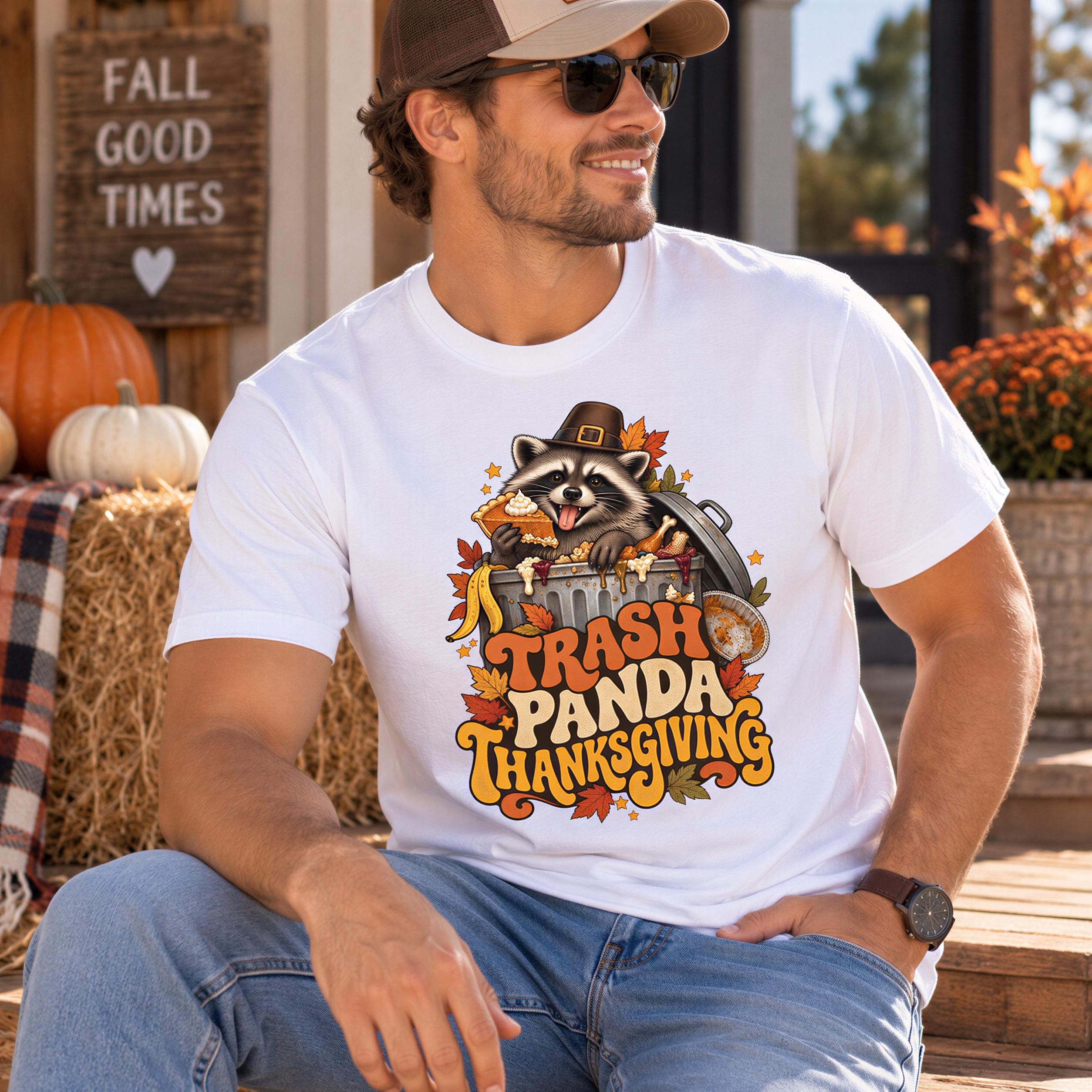 A man wearing a white t-shirt with a graphic design of a raccoon wearing a Thanksgiving hat and holding a trash can, sitting on a wooden bench.