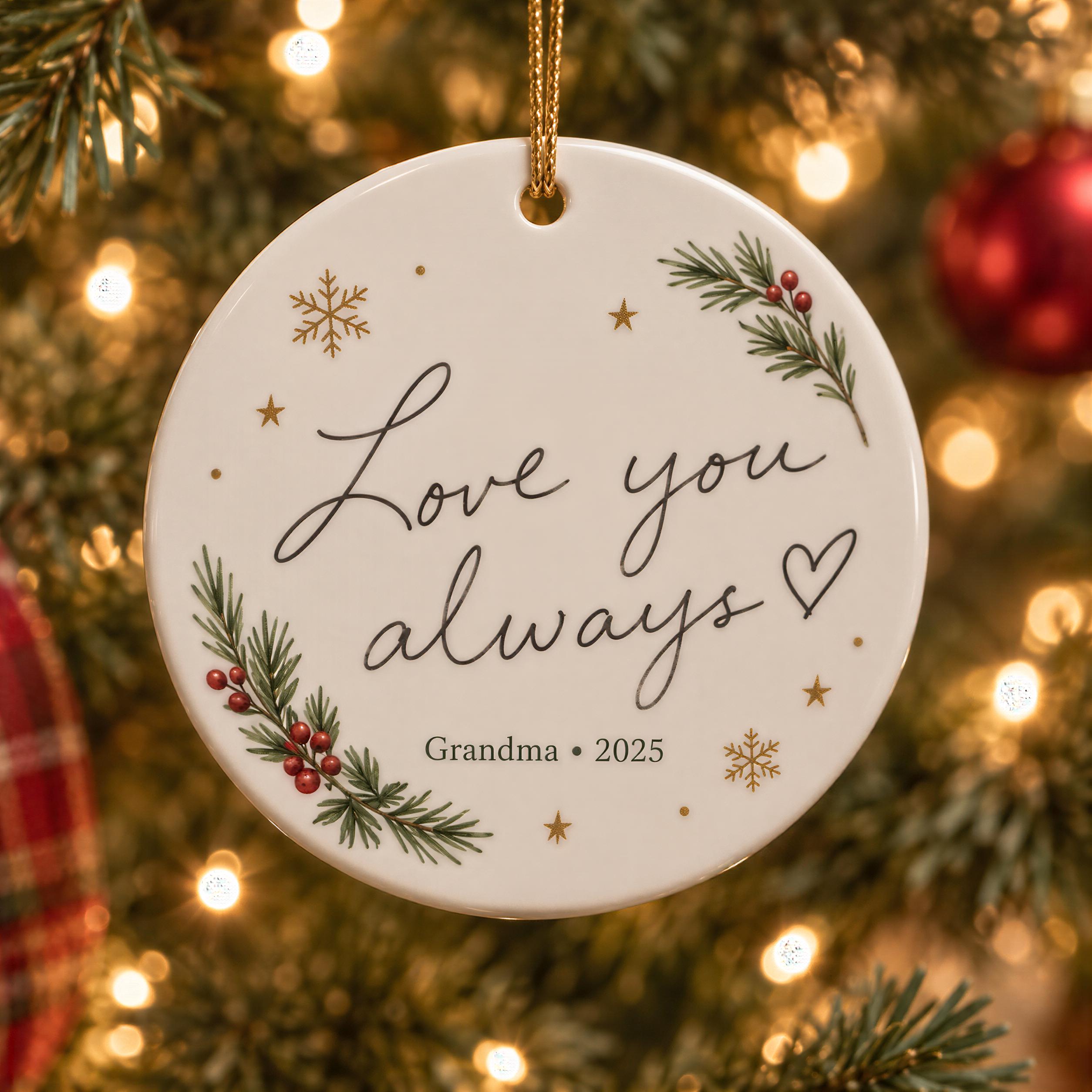 A white ornament with a message that says "Love you always" and a heart symbol, hanging on a Christmas tree with pine branches and red berries.