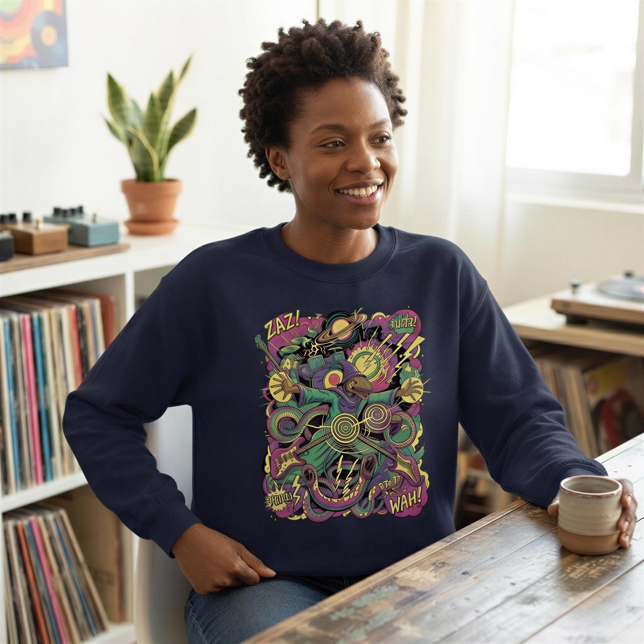 A smiling woman wearing a navy blue sweatshirt with a colorful graphic design on it, sitting at a table and holding a cup.