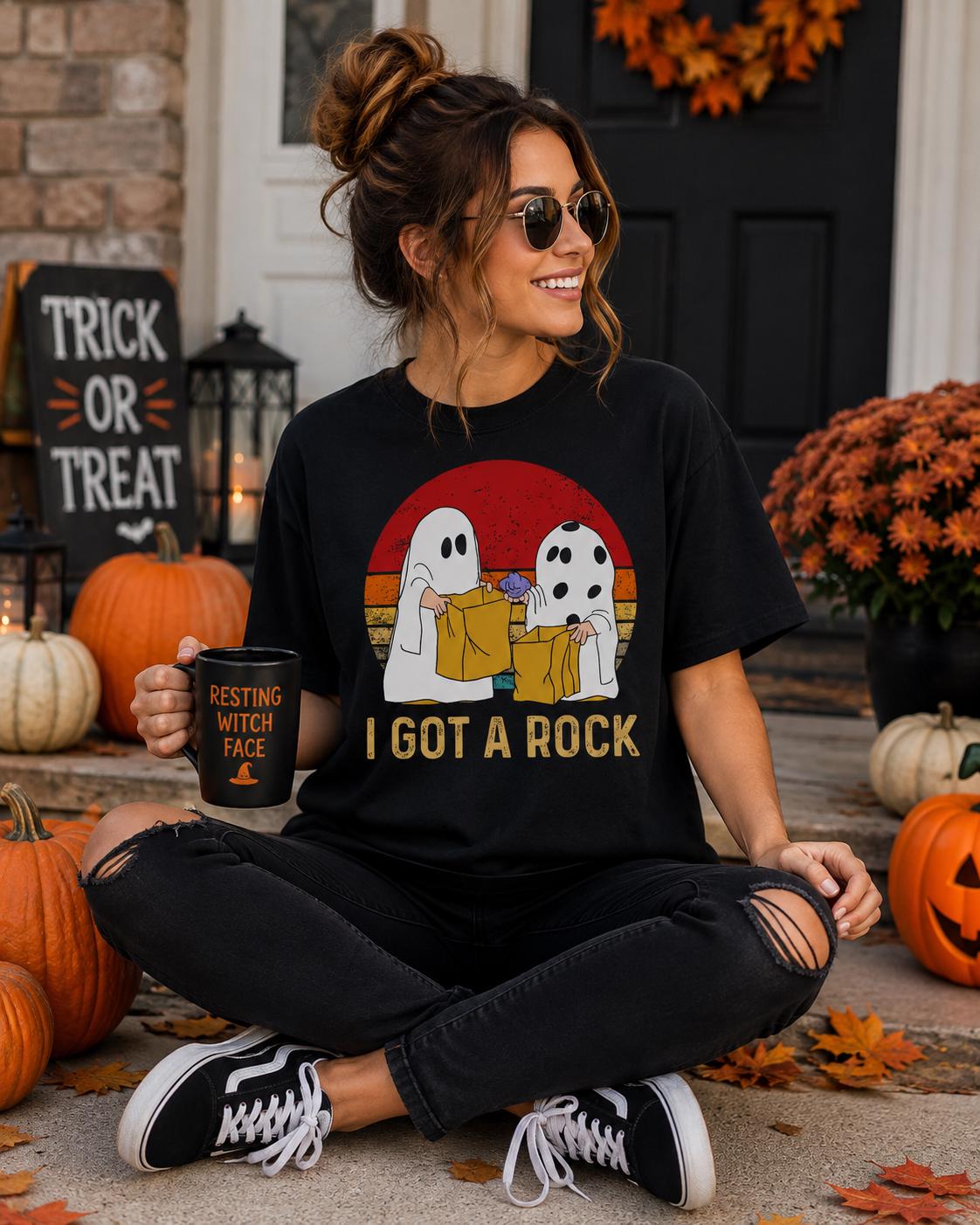 A woman wearing a black t-shirt with a graphic of two ghost characters and the text "I got a rock" is sitting on the ground, holding a mug and surrounded by pumpkins and a Halloween-themed sign.