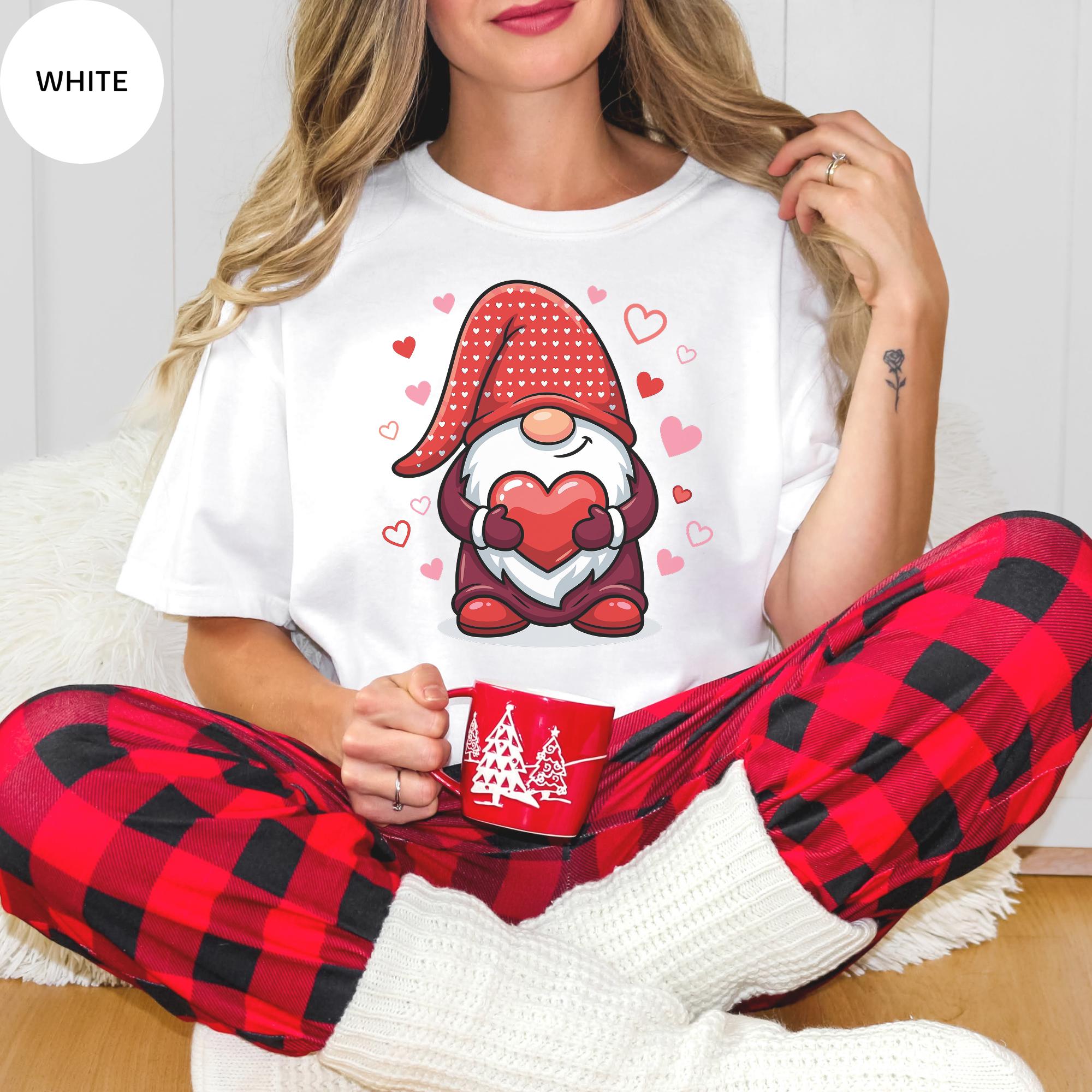 A woman wearing a white t-shirt with a gnome graphic and red heart-shaped hearts, sitting on a white surface and holding a red mug.
