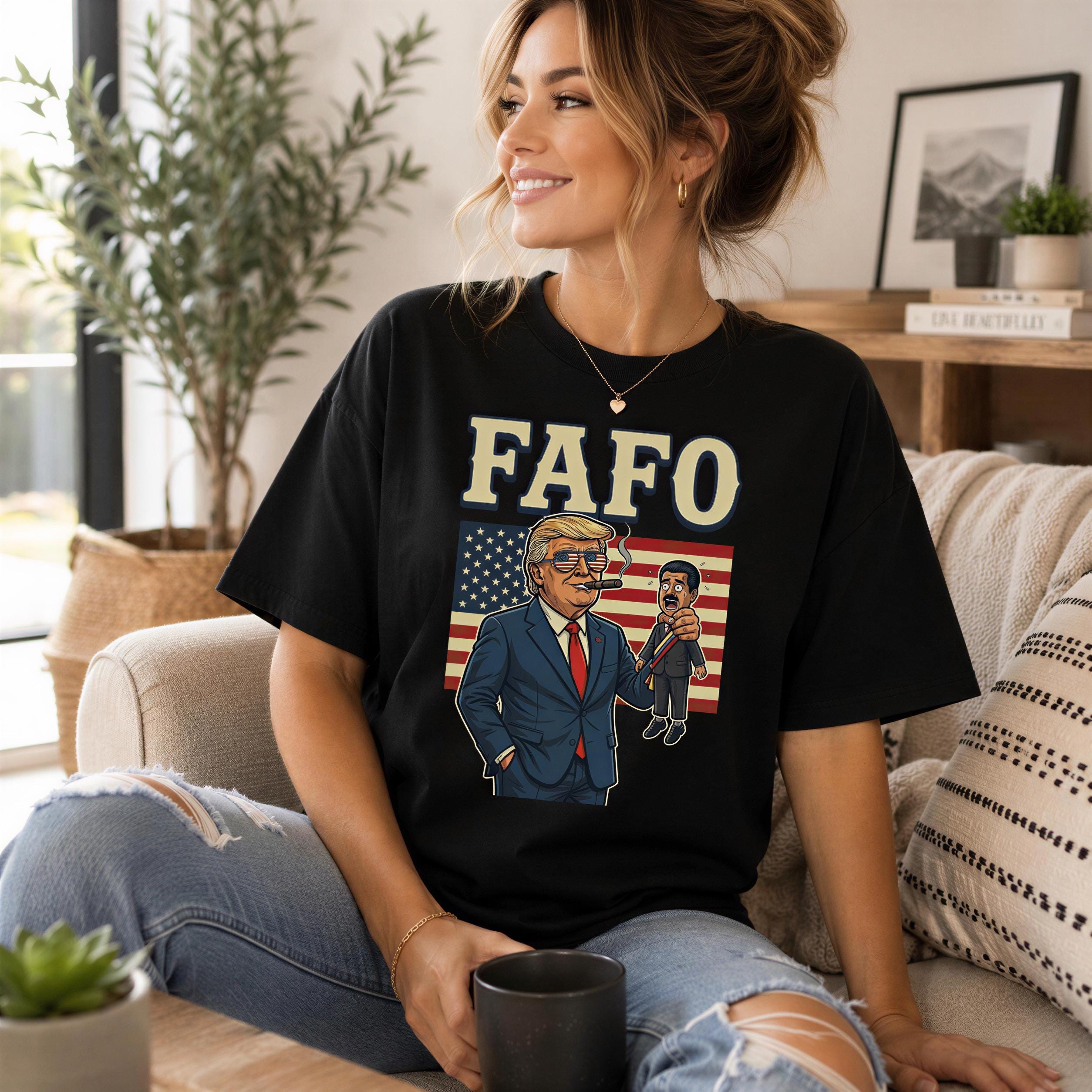A woman wearing a black t-shirt with a graphic of a man in a suit and tie, along with an American flag, is sitting on a couch and smiling.