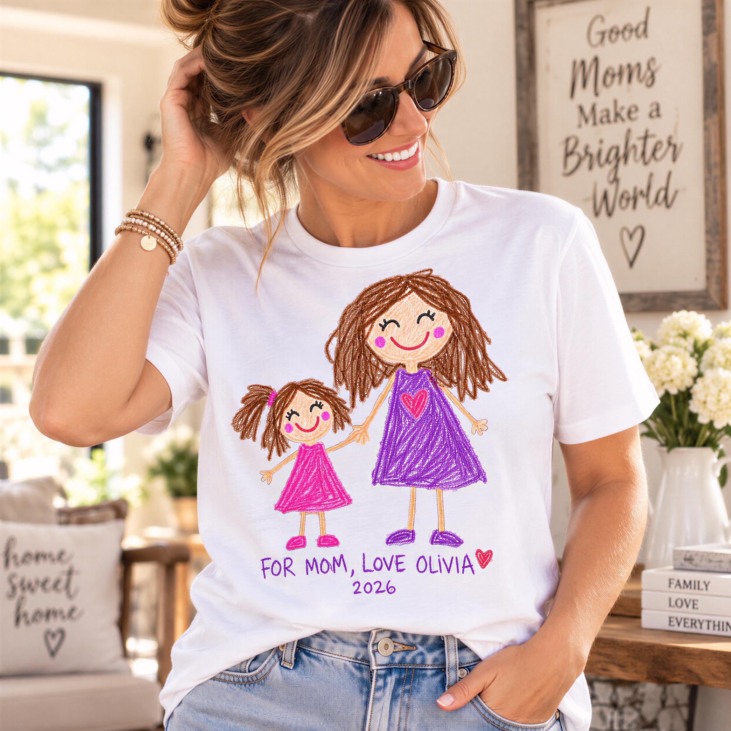 A woman wearing a white t-shirt with a drawing of two girls, one larger and one smaller, and the text "FOR MOM, LOVE OLIVIA 2026" printed on it. She is smiling and adjusting her hair.