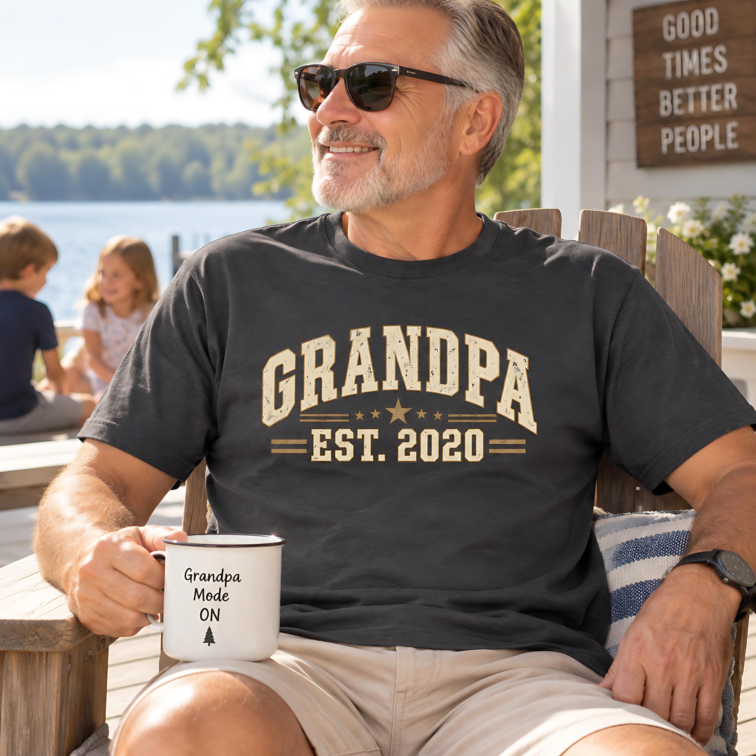 A man wearing a black t-shirt with the text "GRANDPA EST. 2020" is sitting on a bench, holding a white mug with the text "Grandpa mode ON" printed on it.