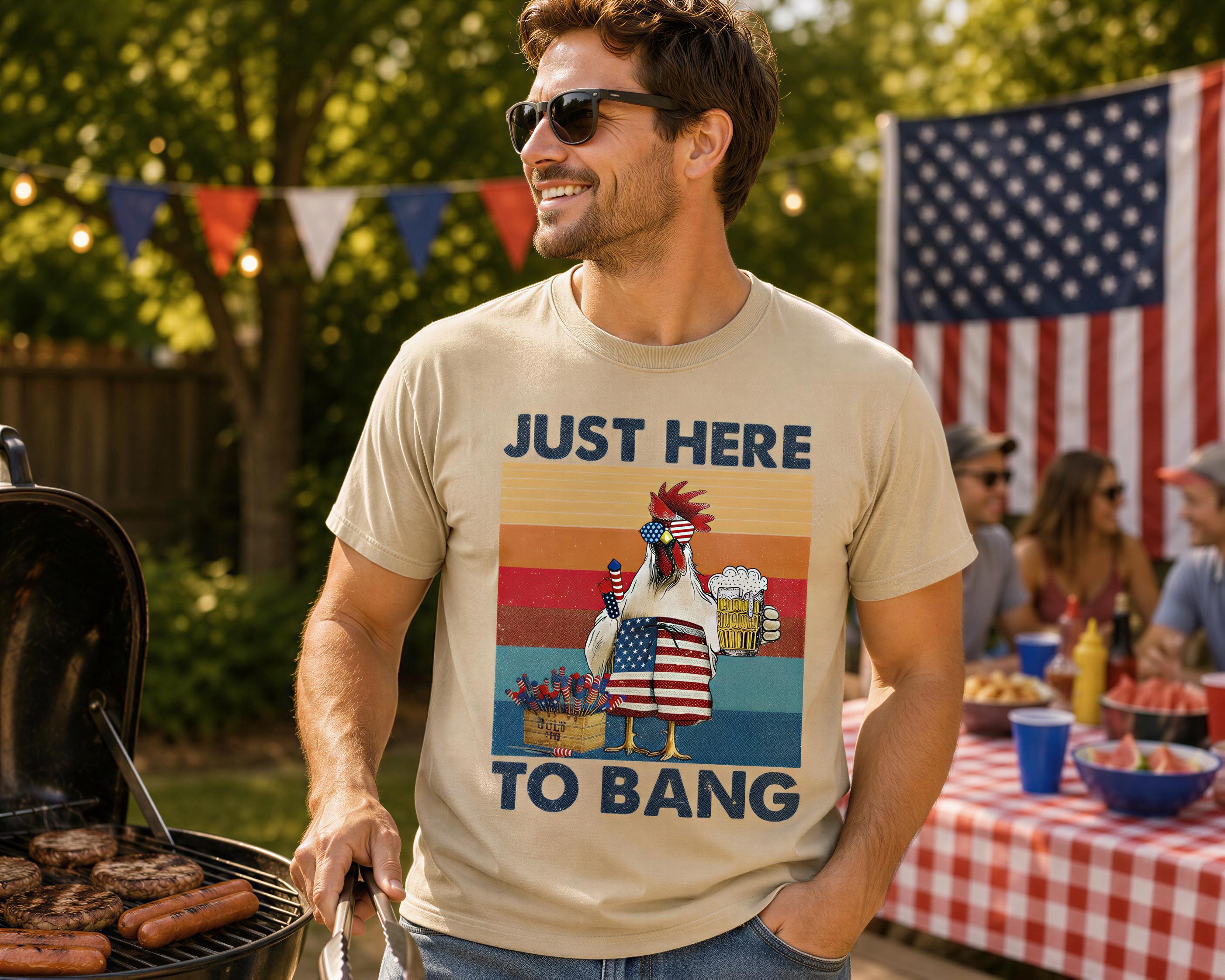 A man wearing a t-shirt with a graphic of a chicken and the text "JUST HERE TO BANG" is standing next to a grill, smiling and holding tongs.