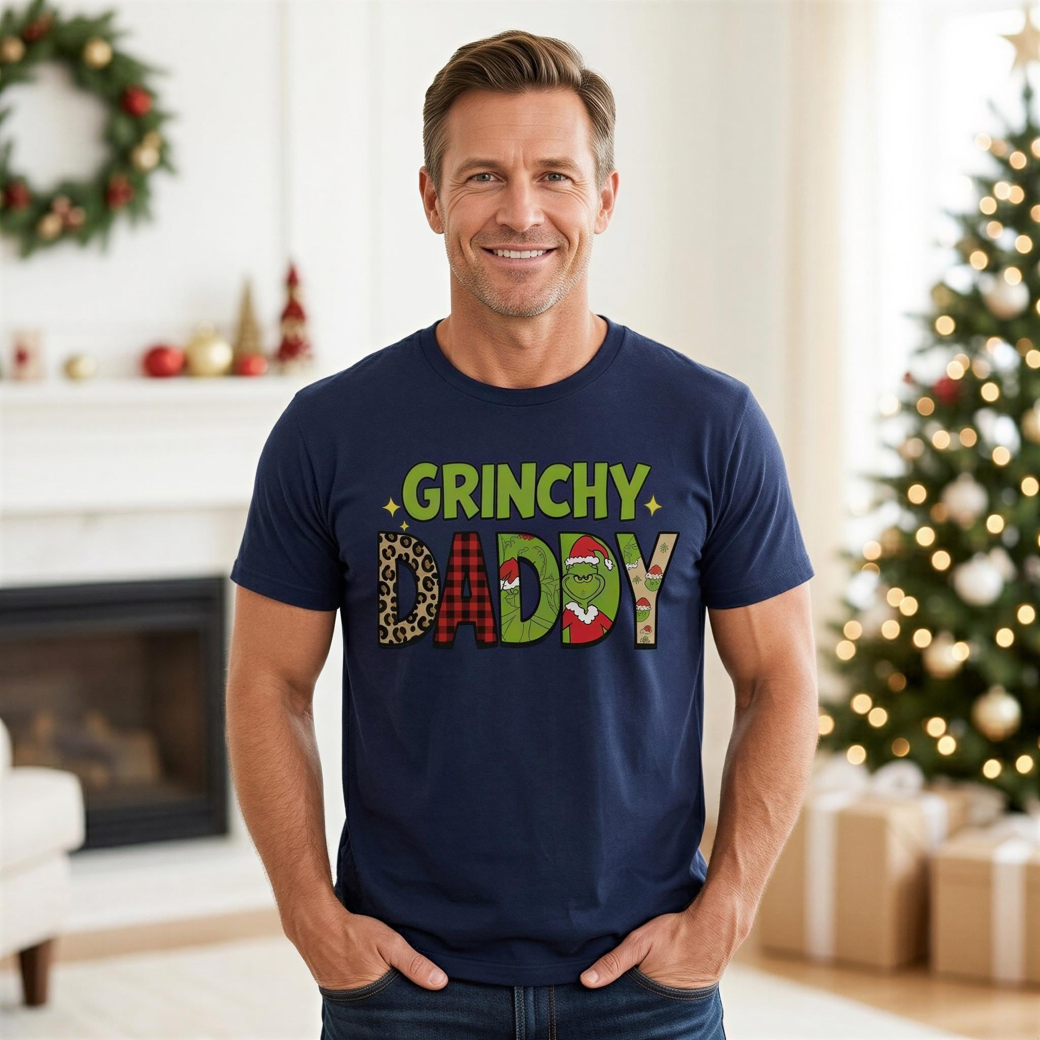 A man wearing a navy blue t-shirt with the text "GRINCHY DADBY" stands in front of a Christmas tree and fireplace.