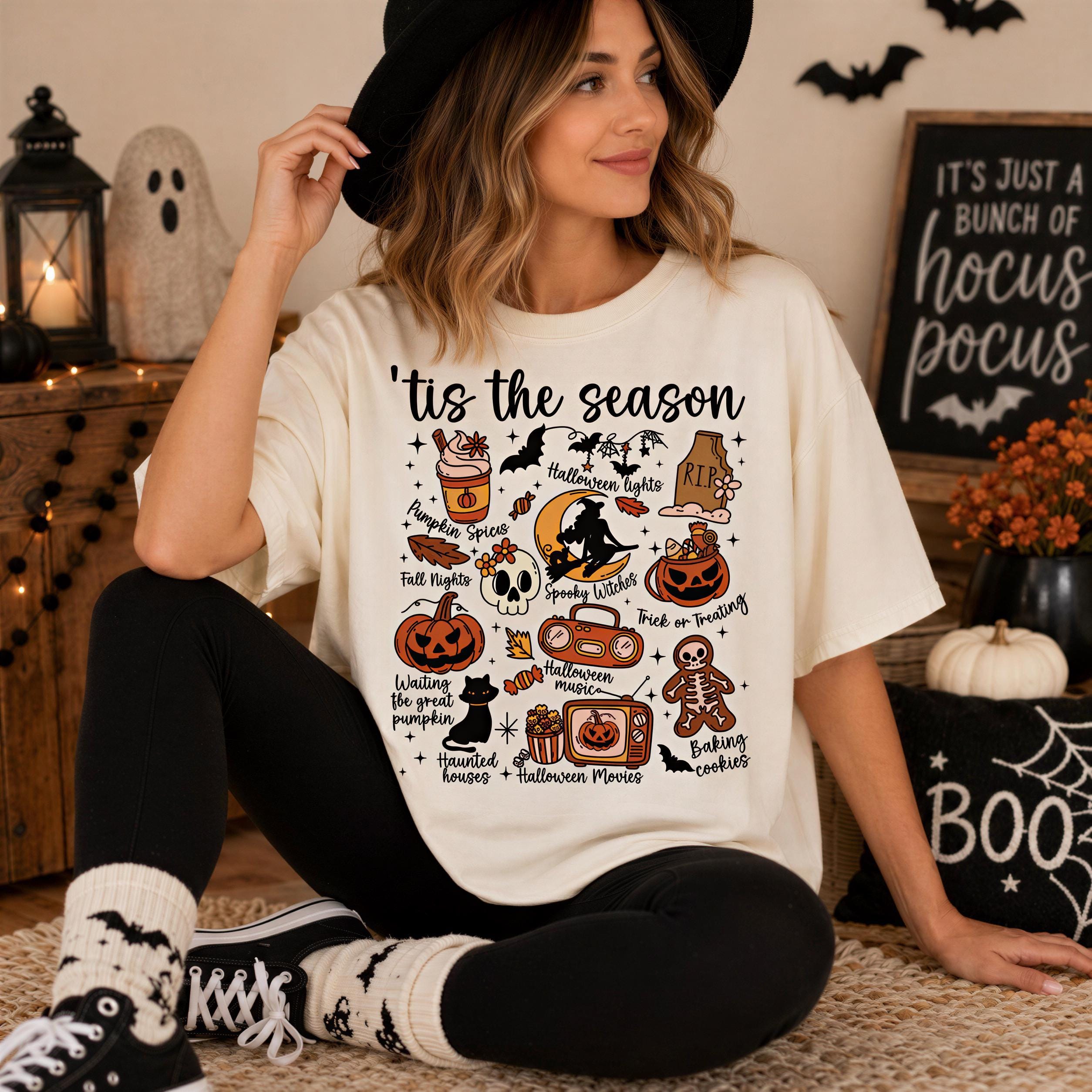 A woman wearing a white t-shirt with a Halloween-themed graphic and text, sitting on a rug in a room decorated for Halloween.