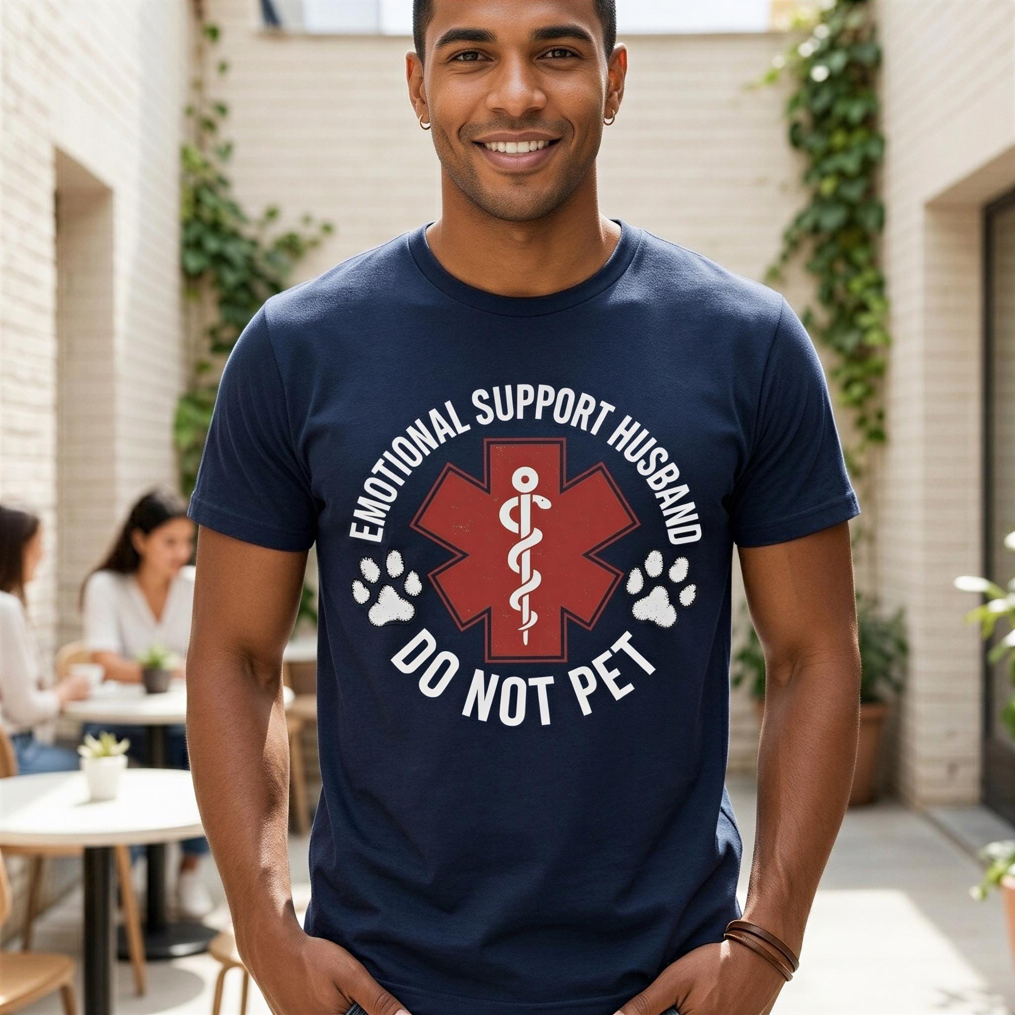 A man wearing a navy blue t-shirt with a red cross symbol and text that says "Emotional Support Husband Do Not Pet" is smiling at the camera.