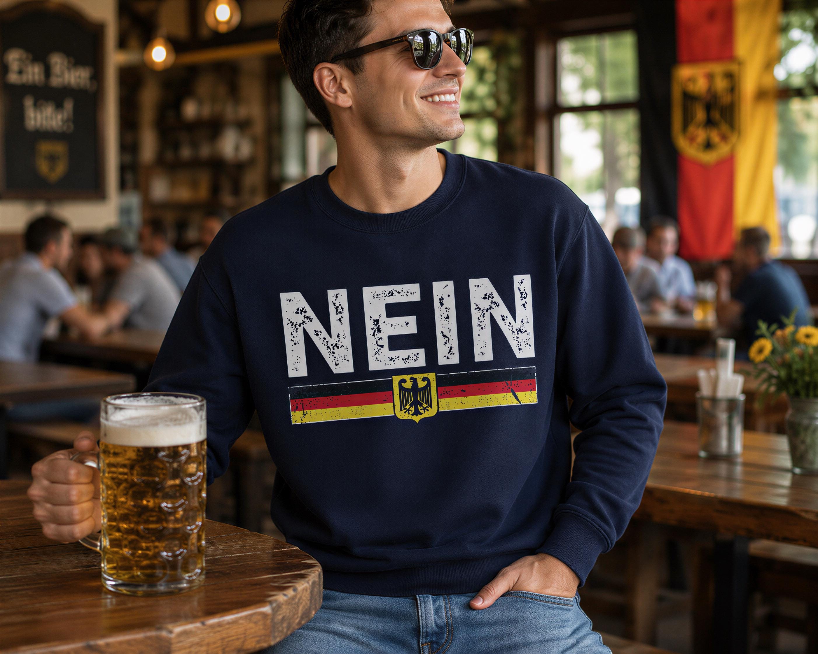 A man wearing a navy blue sweatshirt with the word "NEIN" on it, sitting at a table with a glass of beer in front of him.