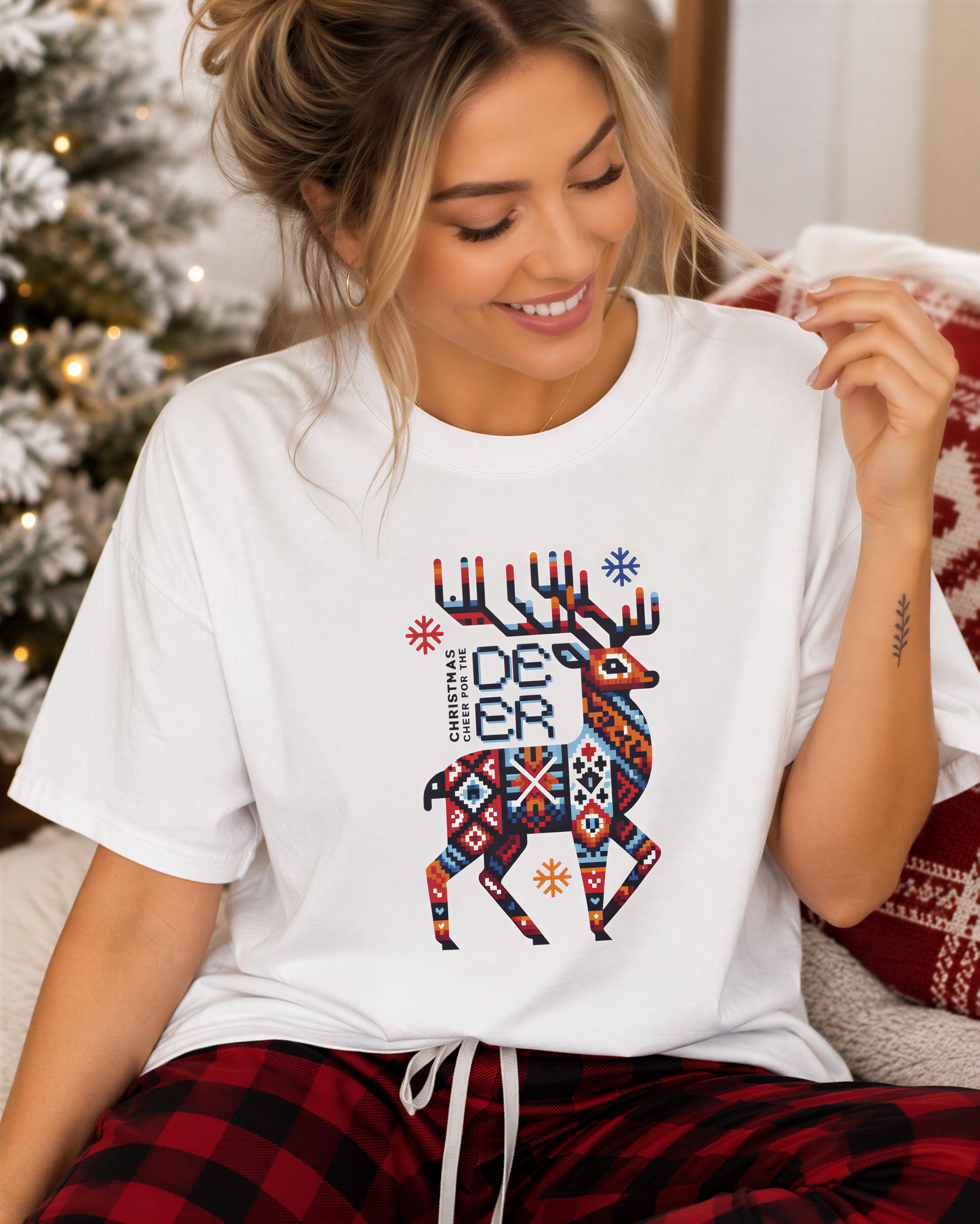 A smiling woman wearing a white t-shirt with a colorful deer graphic and text, sitting on a couch with a Christmas tree in the background.