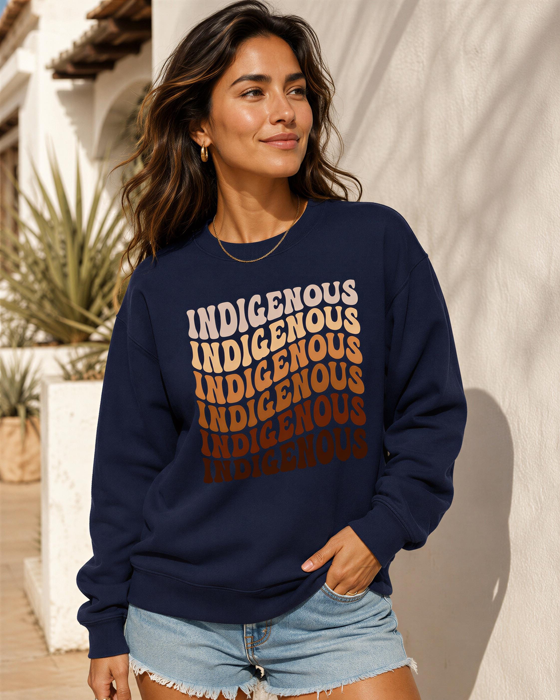 A woman wearing a navy blue sweatshirt with the text "INDIGENOUS INDIGENOUS INDIGENOUS" printed on it, standing in front of a white wall with a plant.