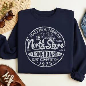 May include: Navy blue sweatshirt featuring a distressed white graphic. The graphic reads "Haleiwa Hawaii 18th Annual North Shore Longboard Surf Competition 1978". The sweatshirt is a casual garment, perfect for surfing enthusiasts.