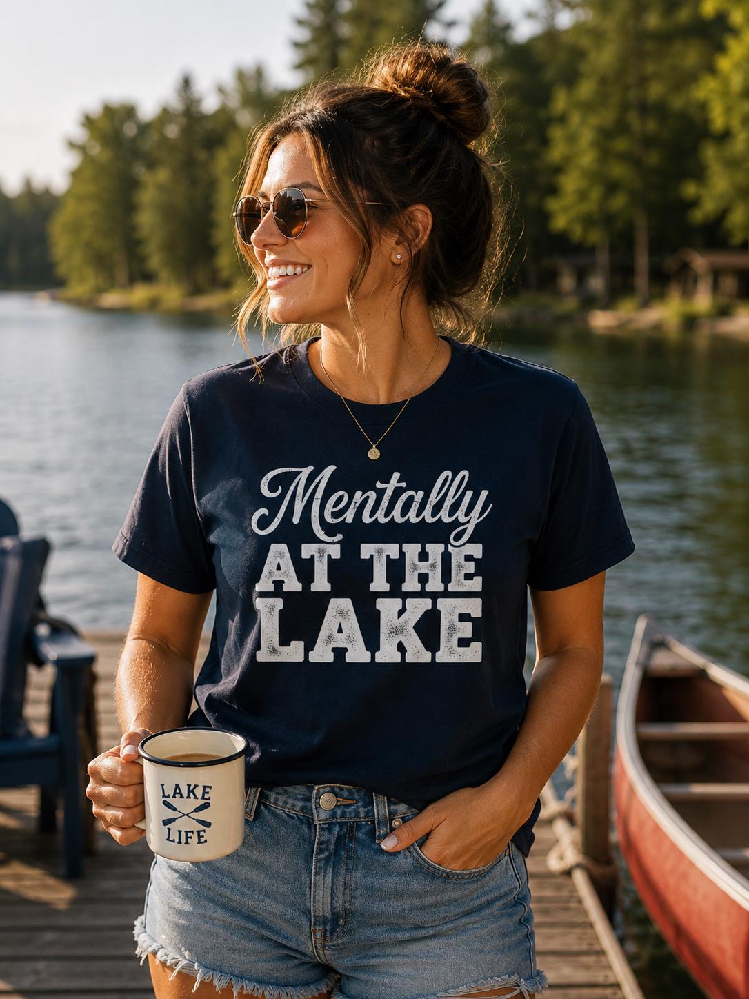 A woman wearing a navy blue t-shirt with the text "Mentally at the Lake" and holding a coffee mug stands near a lake with a boat in the background.