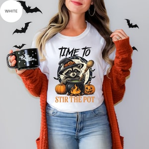 Peut inclure: T-shirt blanc avec un motif sur le thème d'Halloween. Le motif représente un raton laveur portant un chapeau de sorcière remuant un chaudron, avec des citrouilles et le texte "TIME TO STIR THE POT". Des chauves-souris sont en arrière-plan.